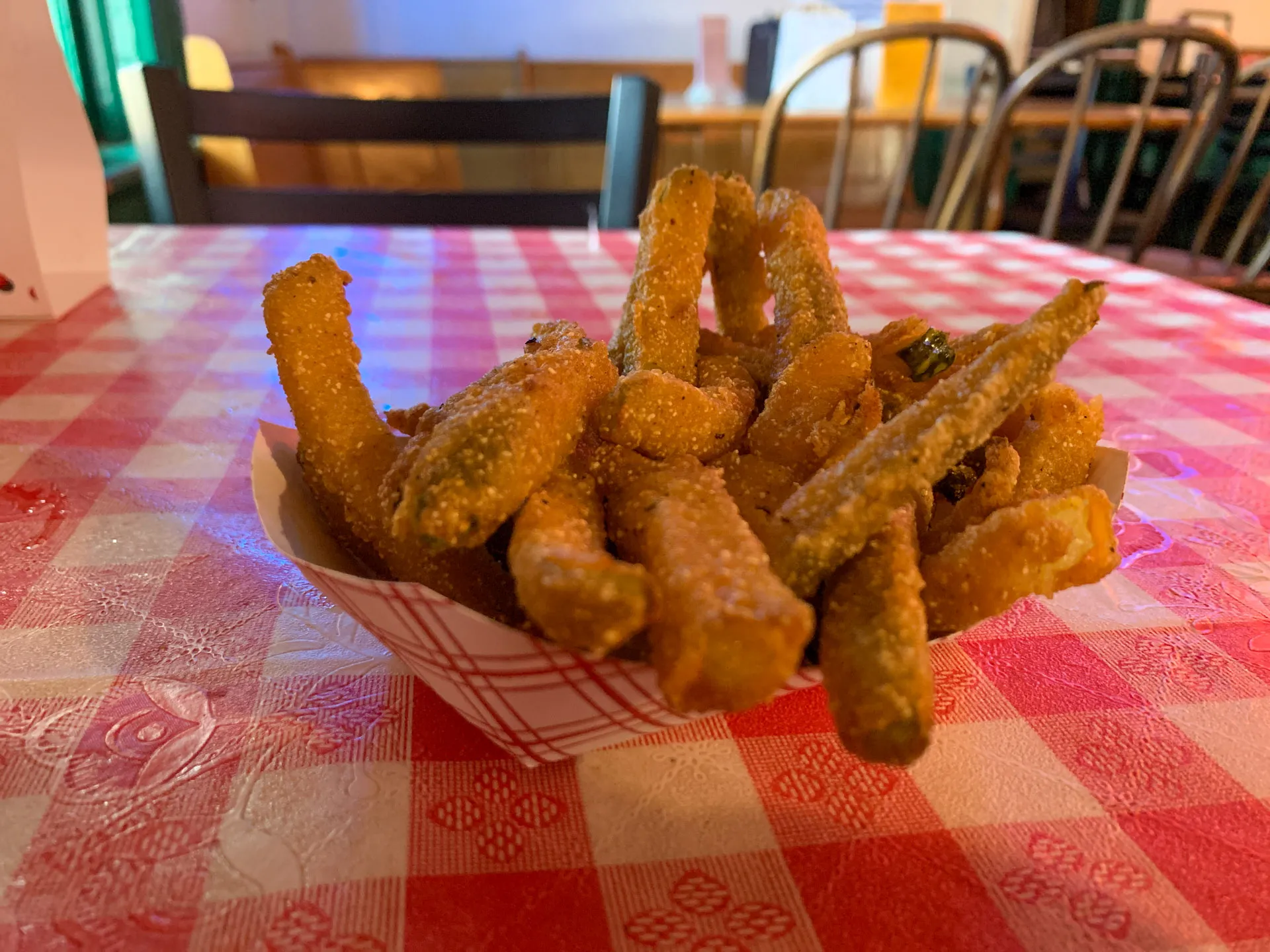 A paper tray filled with deep-fried, breaded pickles sits on a red and white checkered tablecloth. The pickles are golden brown and crispy. In the background, wooden chairs and tables are arranged, creating a cozy dining atmosphere. The lighting is warm, adding to the inviting feel.