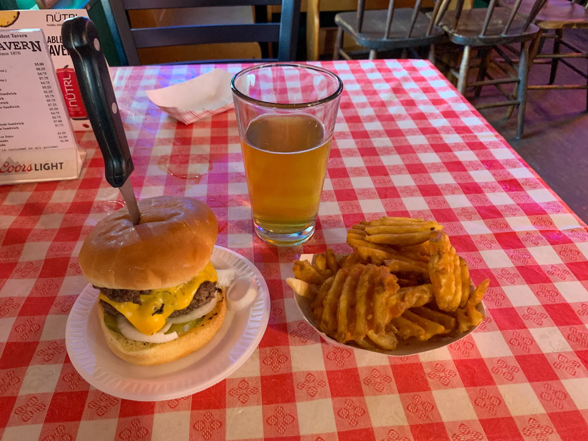 A cheeseburger with a knife through it sits on a white plate on a red and white checkered tablecloth. Next to it are crisscross waffle fries in a bowl and a pint of light amber beer in a glass. A menu and napkin are in the background.