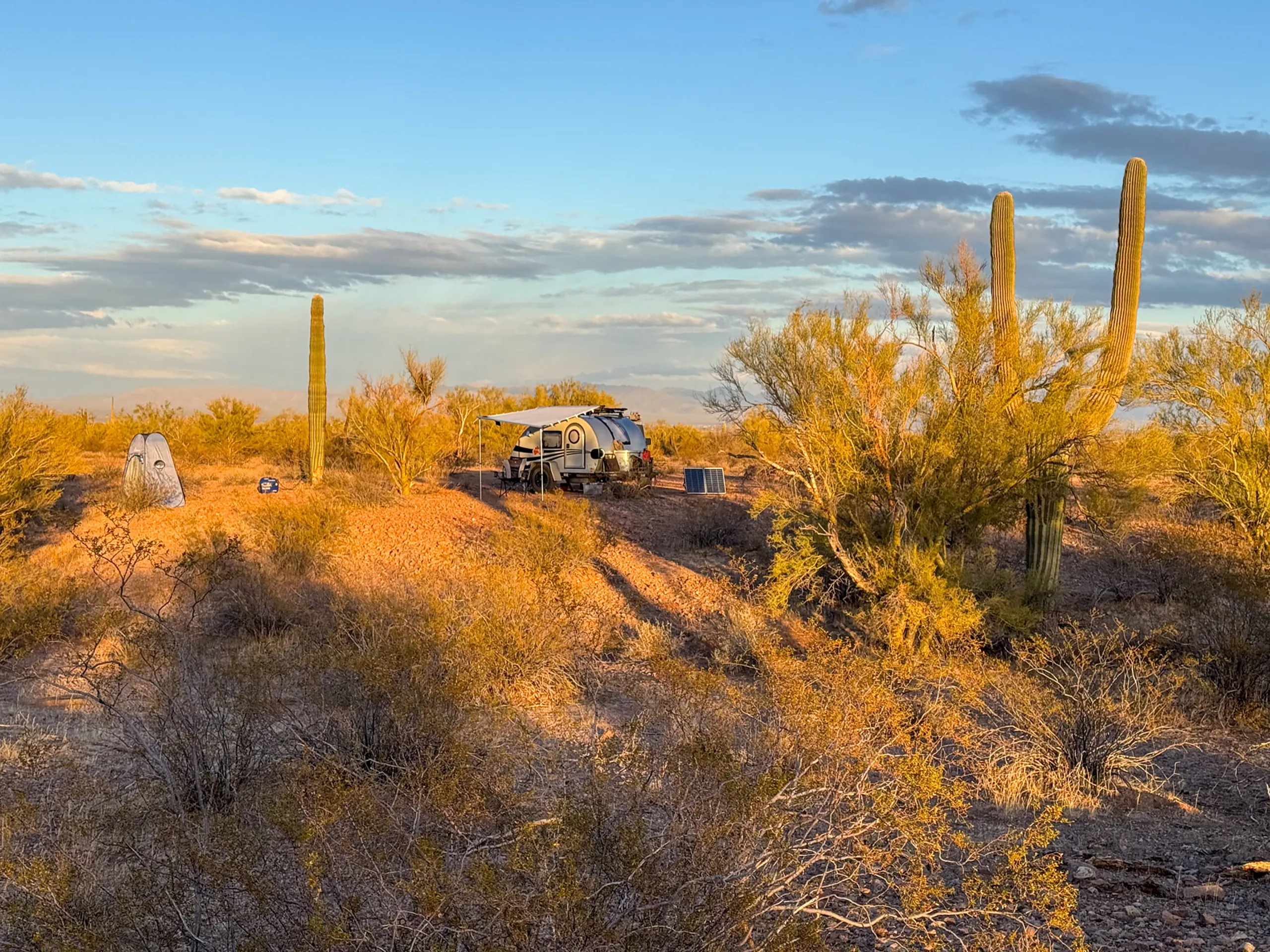 A small camper trailer is parked on a sandy hill in a desert landscape at sunset. Tall cacti and sparse desert shrubs surround the area, with a few camping chairs set up nearby. The sky is a mix of blue and gray with scattered clouds, casting a warm golden light on the scene.