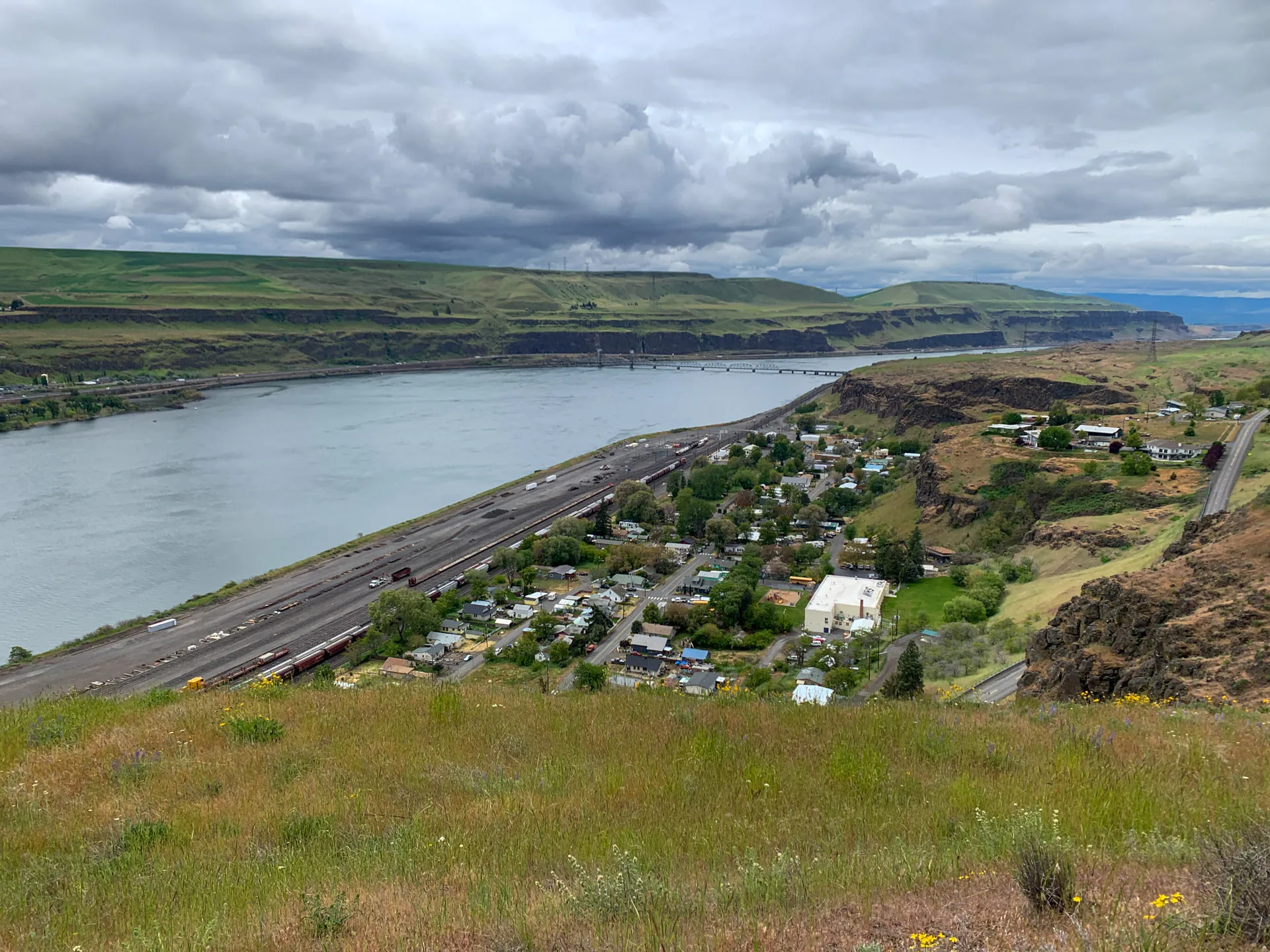 View of a river with a bridge in the distance, surrounded by grassy hills and small settlements. The sky is cloudy, casting soft shadows over the landscape. A road runs parallel to the river, with scattered houses nestled among green fields and rocky terrain.