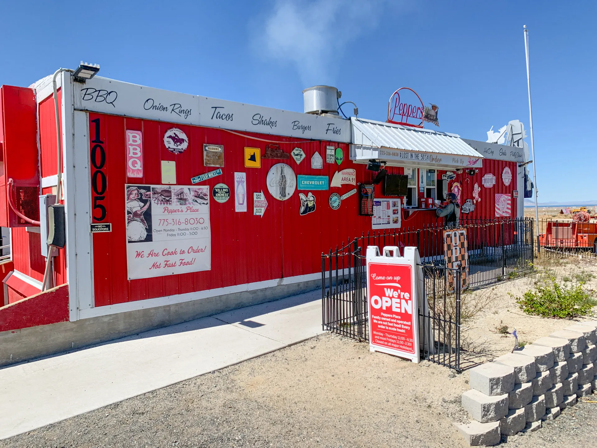 A red food trailer with various stickers and signs offers BBQ, onion rings, tacos, shakes, burgers, and fries. A sign with the phone number is displayed. A "We're Open" sign is on a stand in front, beside a small brick wall and a metal fence, under a clear blue sky.