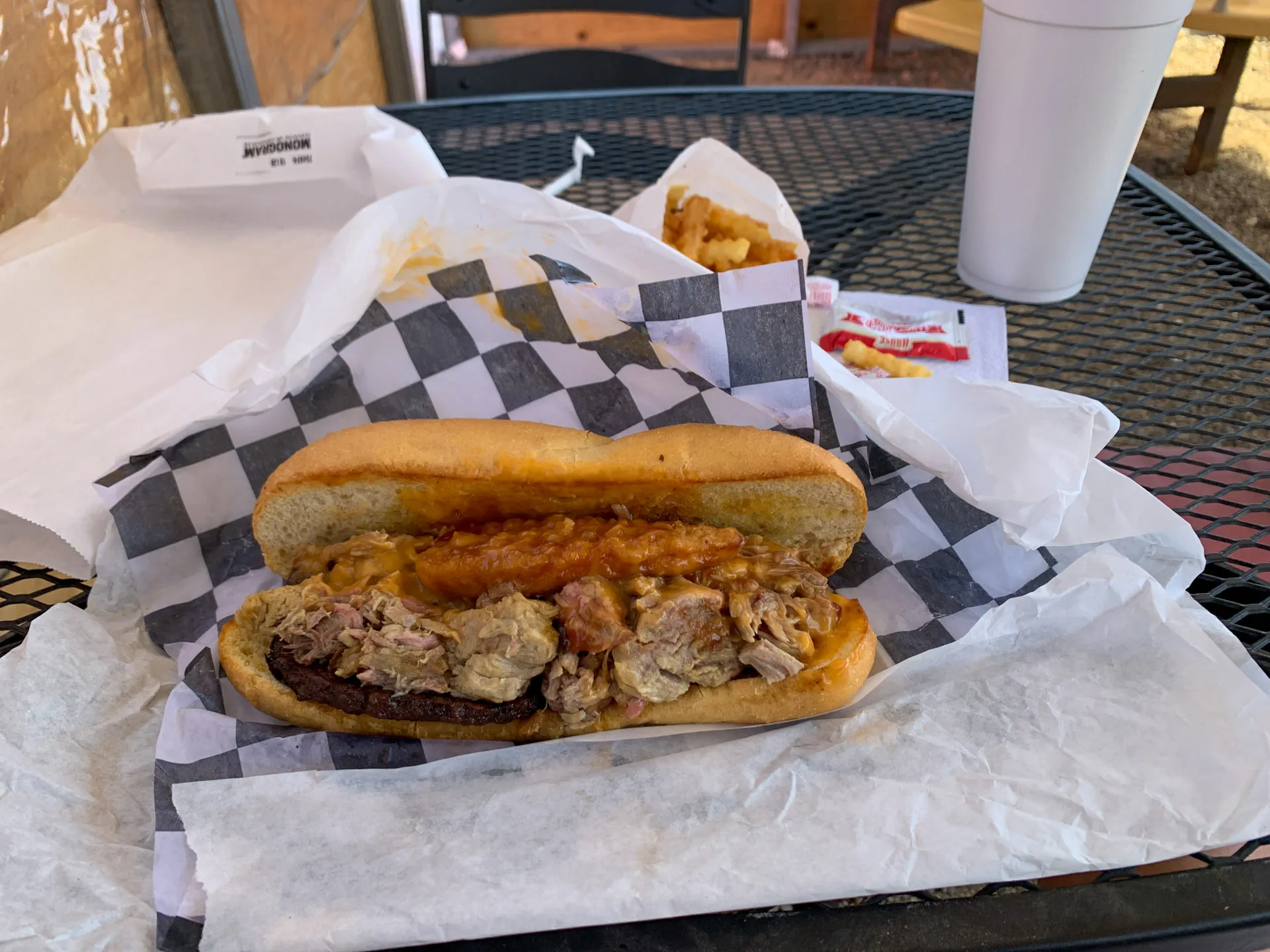 A hearty sandwich with pulled pork, a burger, a chicken tender, and sauce on a checkered paper-lined table. In the background, a small tray of fries with a ketchup packet sits beside a white cup. The setting appears to be an outdoor table with a metal mesh surface.
