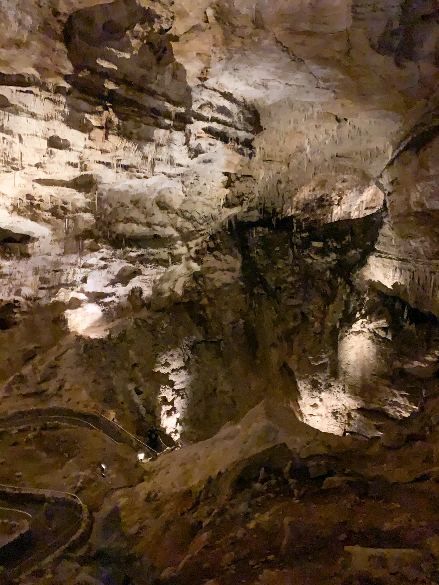A dimly lit cave interior features rugged rock formations with stalactites hanging from the ceiling. The walls display varied textures, illuminated by soft lighting. A winding path or walkway is visible along the cave floor, leading deeper into the cavernous space.