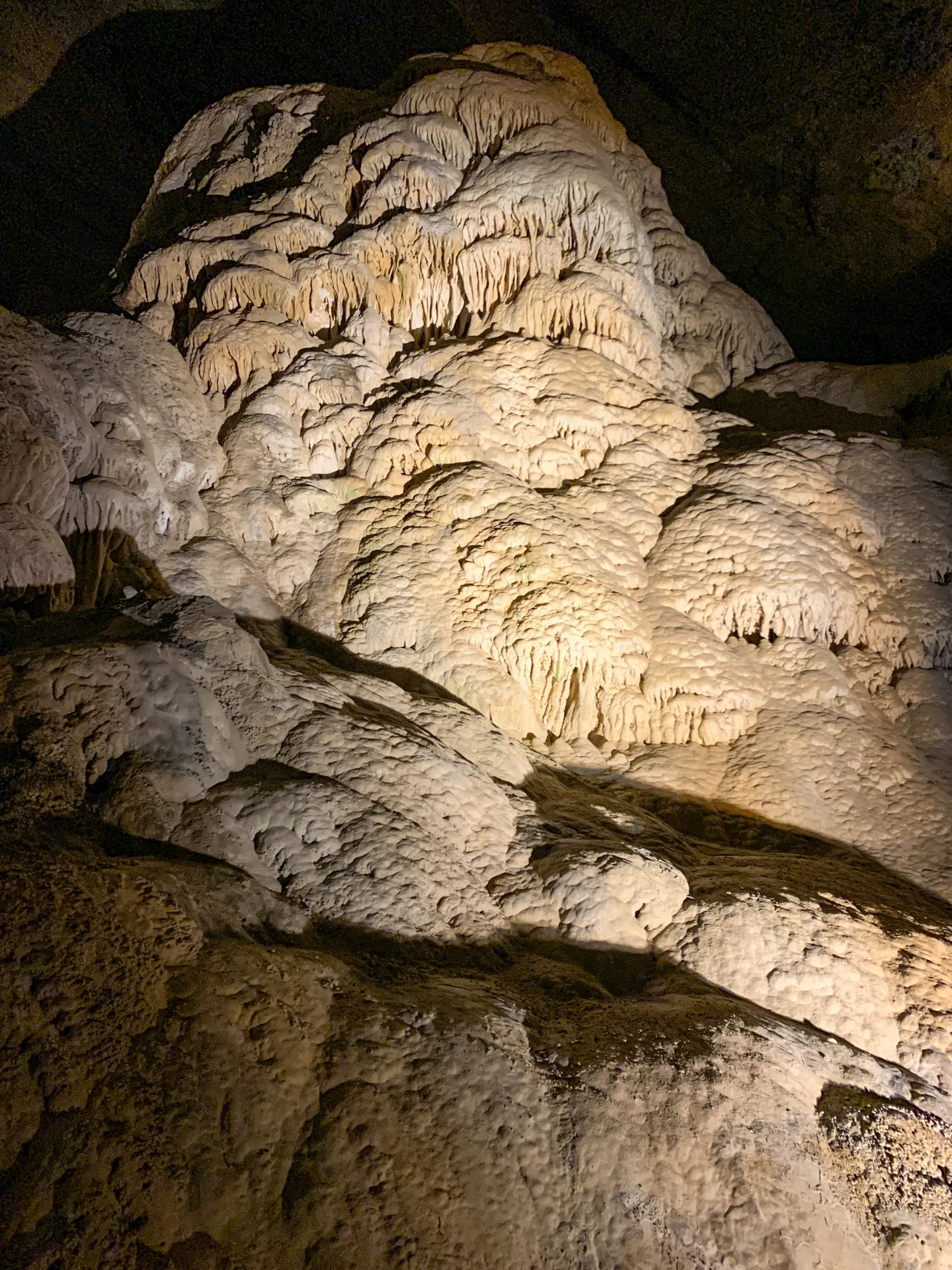 Illuminated stalagmite formation inside a dimly lit cave, showcasing textured, beige rock surfaces with rounded, wavy shapes. The cave walls are dark, providing contrast to the highlighted formation, which features small stalactites hanging from its top edges.