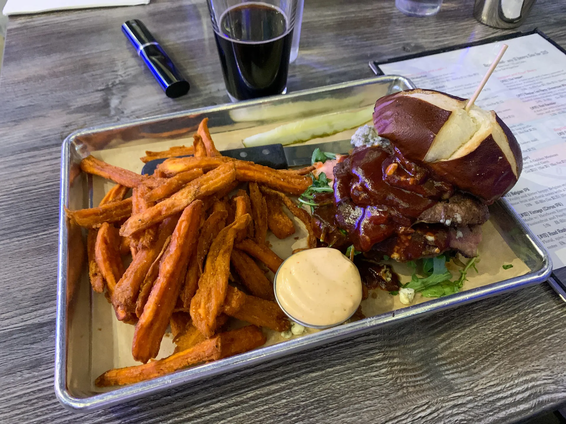 A metal tray on a wooden table holds a sandwich with a pretzel bun, lettuce, and sauce beside a pile of sweet potato fries. A pickle spear and a small dish of dipping sauce accompany the meal. A glass of dark beverage and a menu are in the background.