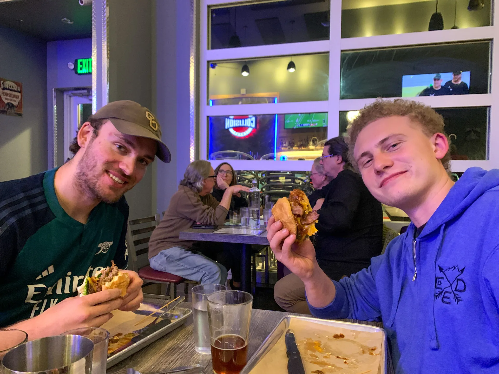 Two people are sitting at a restaurant table, each holding a burger. Both are smiling at the camera. The person on the left is wearing a green and black shirt with a cap, and the one on the right is in a blue hoodie. Behind them, other diners sit at tables. The setting appears casual.