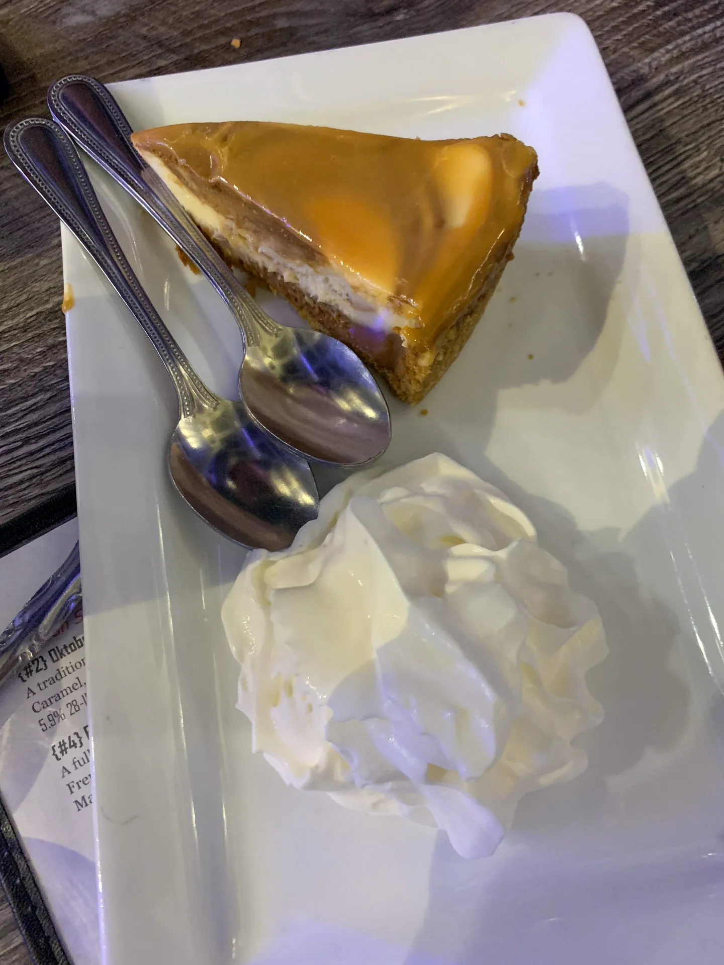 A slice of caramel cheesecake on a white rectangular plate with a caramel glaze on top. Two small metal spoons rest beside it. A generous dollop of whipped cream is placed on the plate beside the cheesecake. The plate is on a wooden table.
