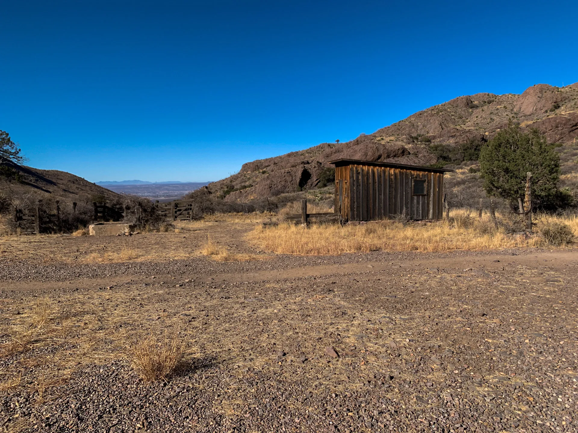 A rustic wooden shed stands in a dry, barren landscape under a clear blue sky. The ground is covered with scattered shrubs and gravel. In the background, rocky hills and sparse trees are visible. A distant view of the horizon is seen between the hills.
