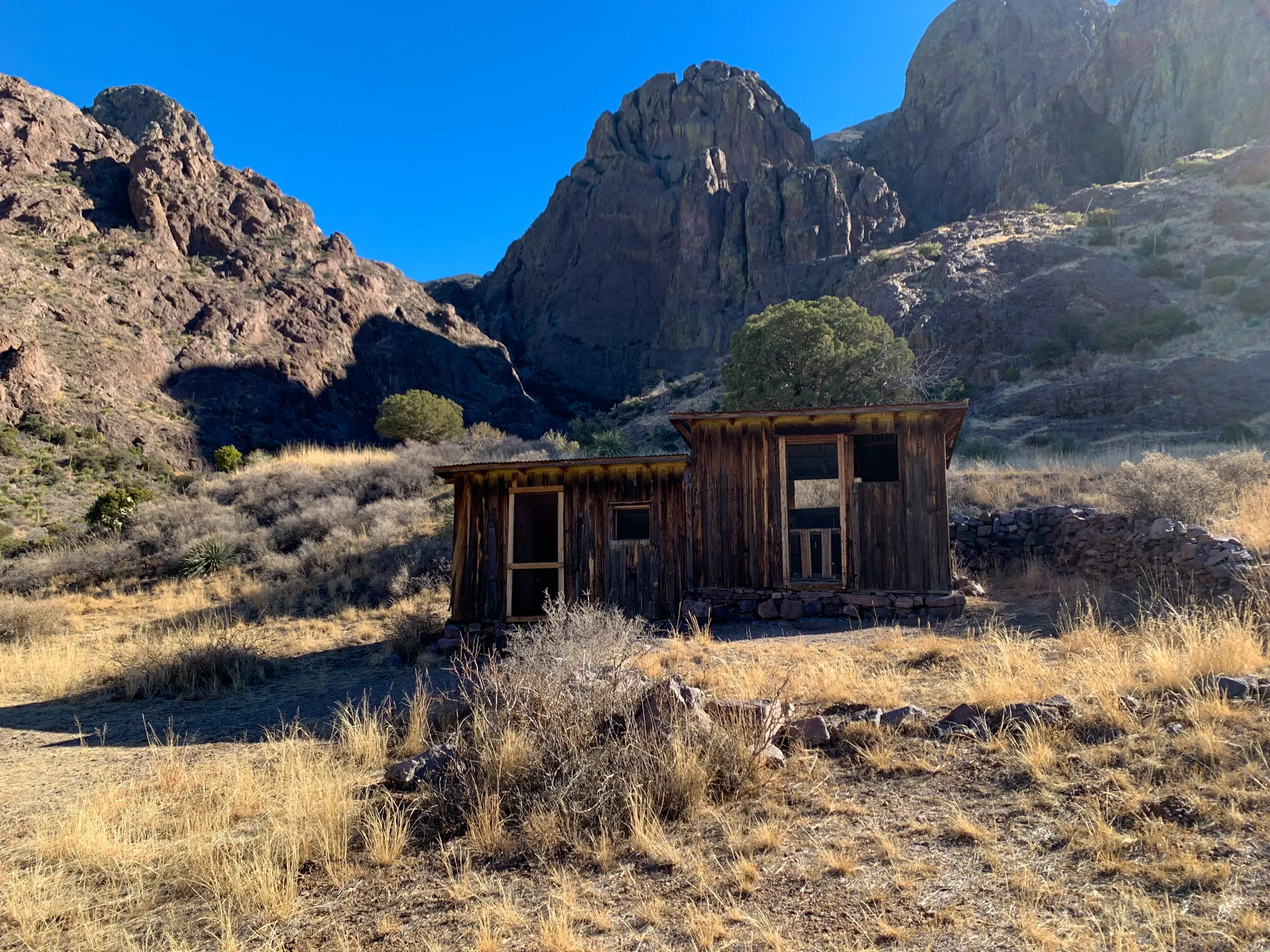 Wooden shacks stand in an arid landscape with dry grass and sparse bushes, set against a backdrop of rugged, sunlit mountains and a clear blue sky. The structures appear weathered, suggesting age and abandonment. Shadows create contrast on the mountainous terrain.