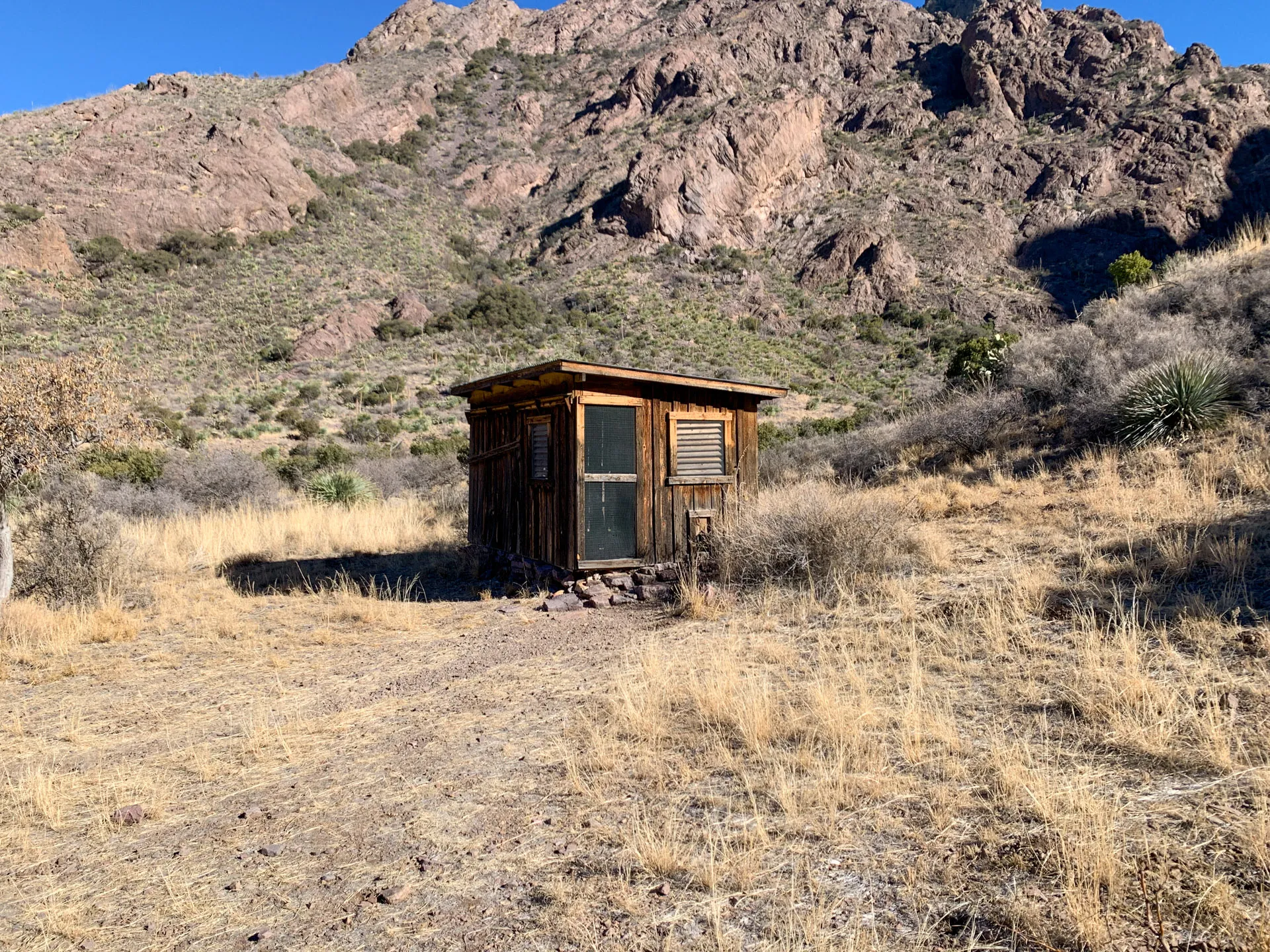 A small wooden shack with a metal roof and a door stands in a dry, grassy field surrounded by brush. In the background, there is a rocky hillside under a clear blue sky. The landscape is arid, with scattered vegetation typical of a desert environment.
