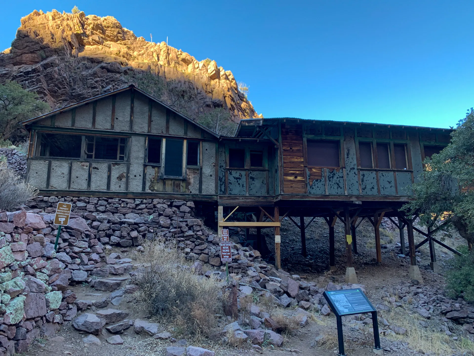 A rustic, weathered wooden cabin elevated on stilts in a rocky desert landscape. The surrounding terrain has dry vegetation and stones. There's an informational sign in front, and a rocky hill bathed in golden sunlight rises behind the cabin. A clear blue sky is above.