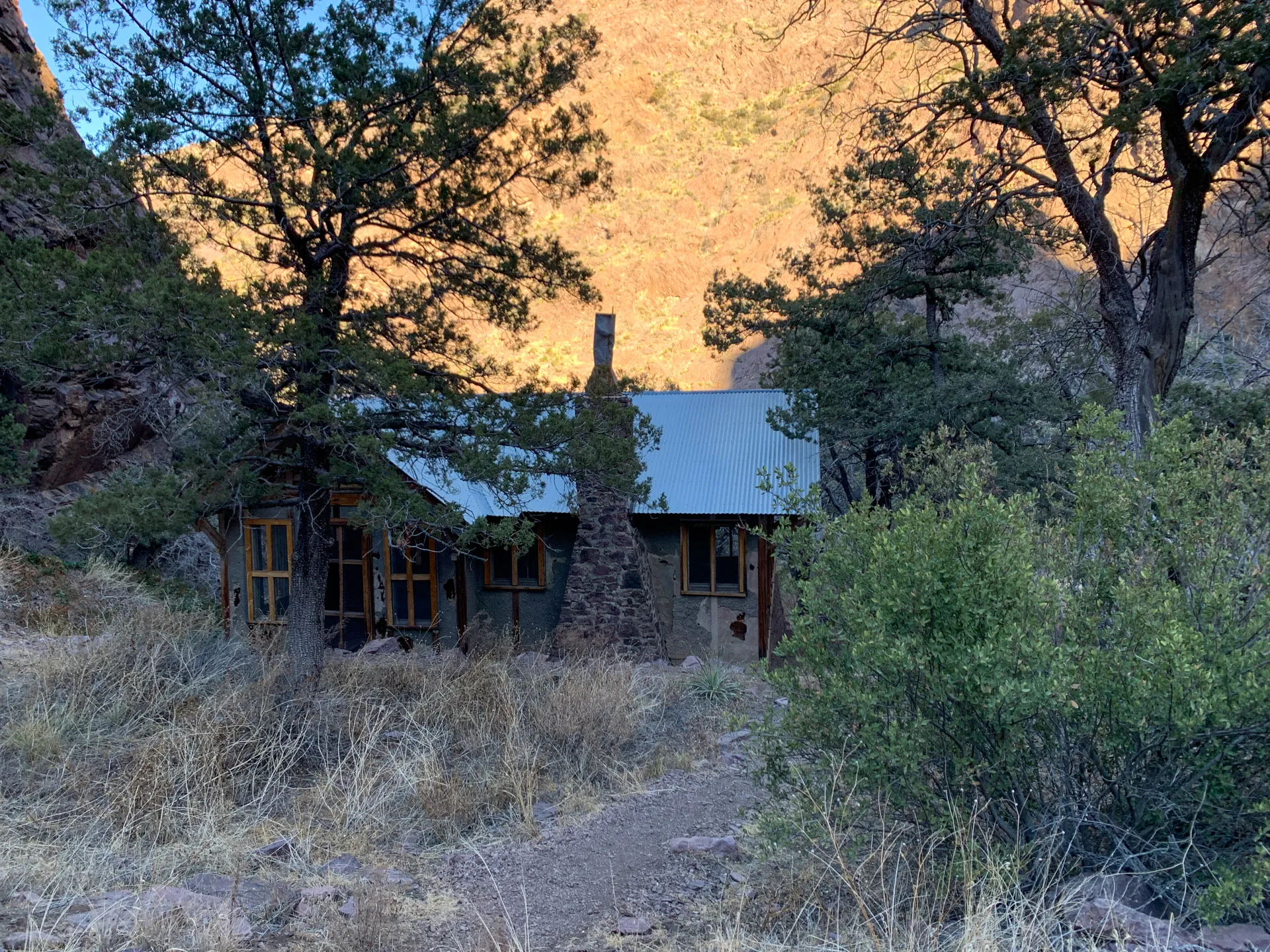 A small, rustic cabin with a metal roof nestled among pine trees in a mountainous area. The cabin has stone and wood construction, with a chimney and several windows. Surrounding it are dry grass and bushes. The background showcases sunlit rocky cliffs under a clear sky.