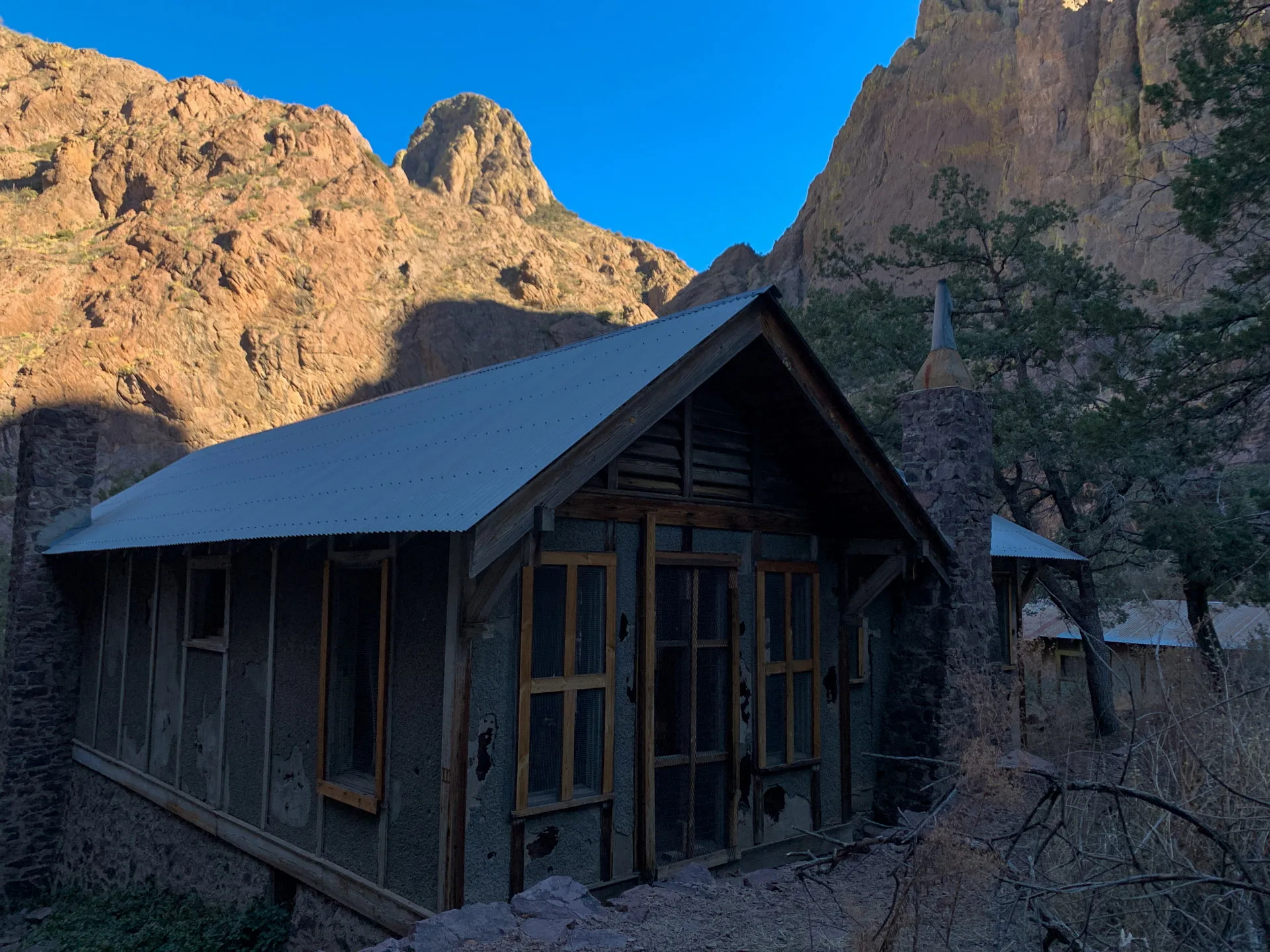 An old, rustic cabin with a metal roof nestled in a rocky canyon. The structure has wooden beams and glass windows, partially shaded by a tree. The backdrop features rugged, sunlit rock formations under a clear blue sky. Sparse vegetation surrounds the cabin.