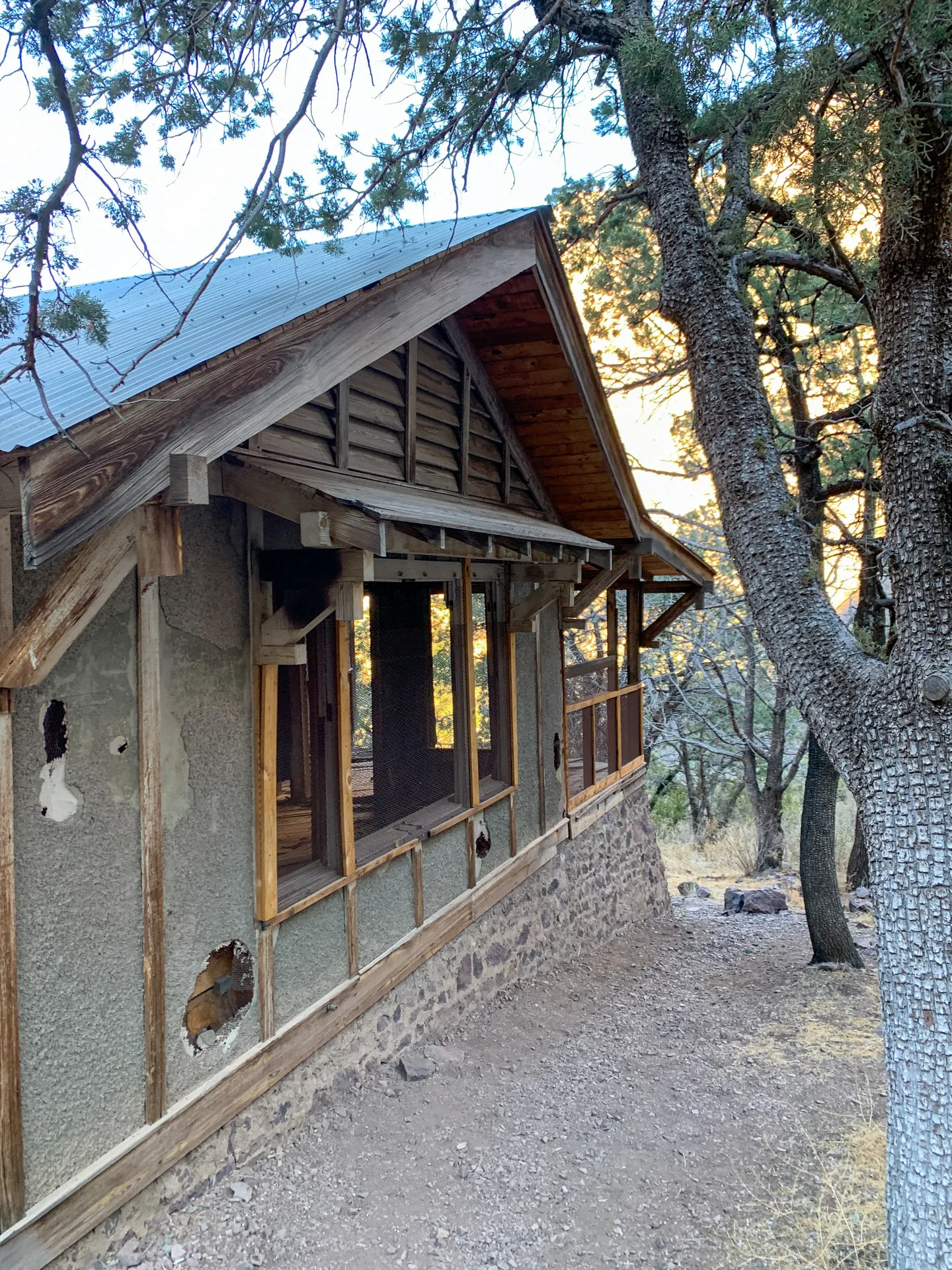 Abandoned wooden cabin with visible wear, missing windows, and damaged walls. The structure sits on a stone foundation amid a wooded area. Sunlight filters through the trees, casting dappled shadows on the ground, hinting at a quiet, serene forest setting.