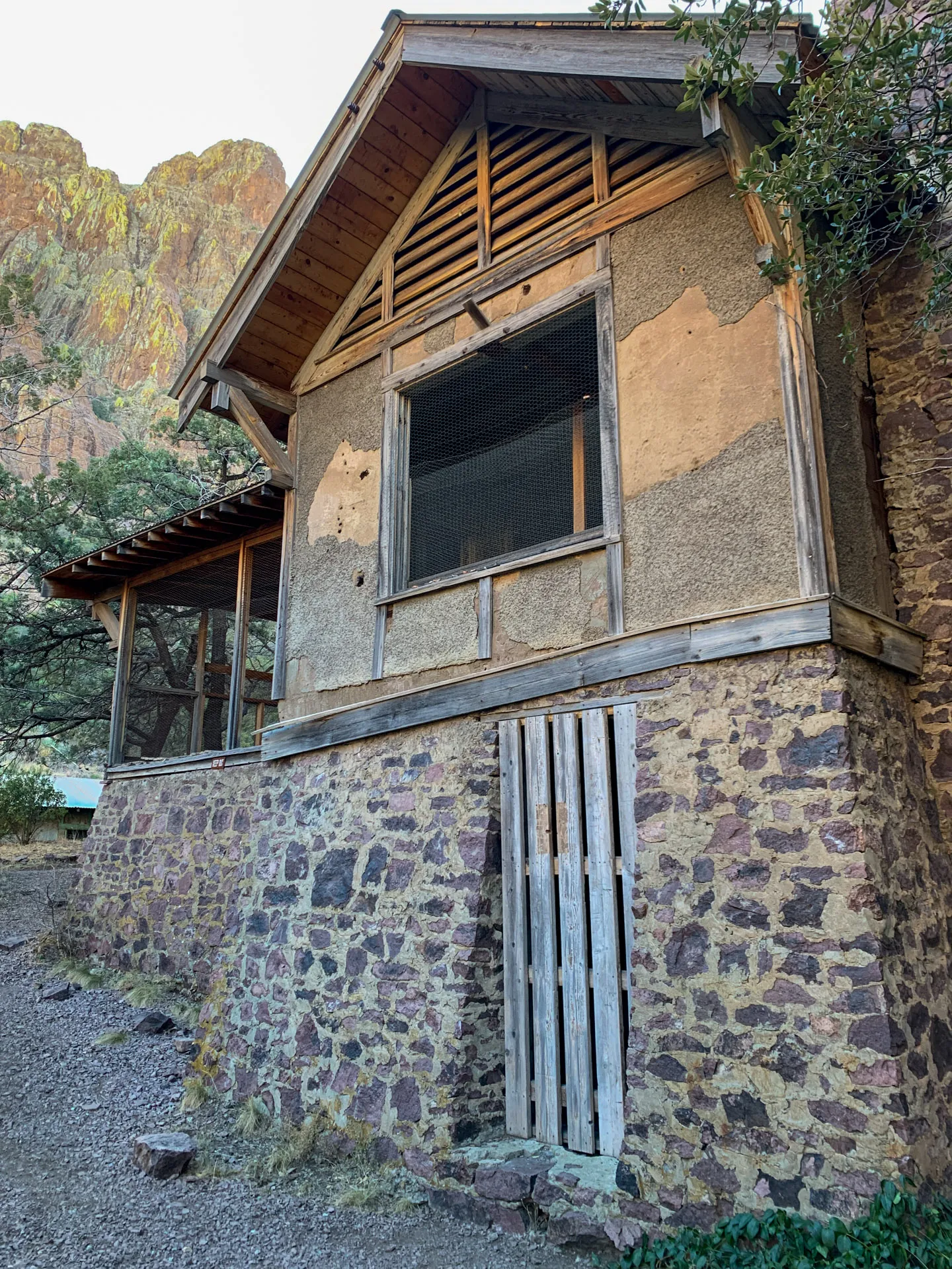 A rustic building with a stone foundation, wooden siding, and a slanted roof. The front features large screened windows and wooden slats. A tree and rocky terrain are visible around it. In the background, there's a sunlit cliff with shrubs.