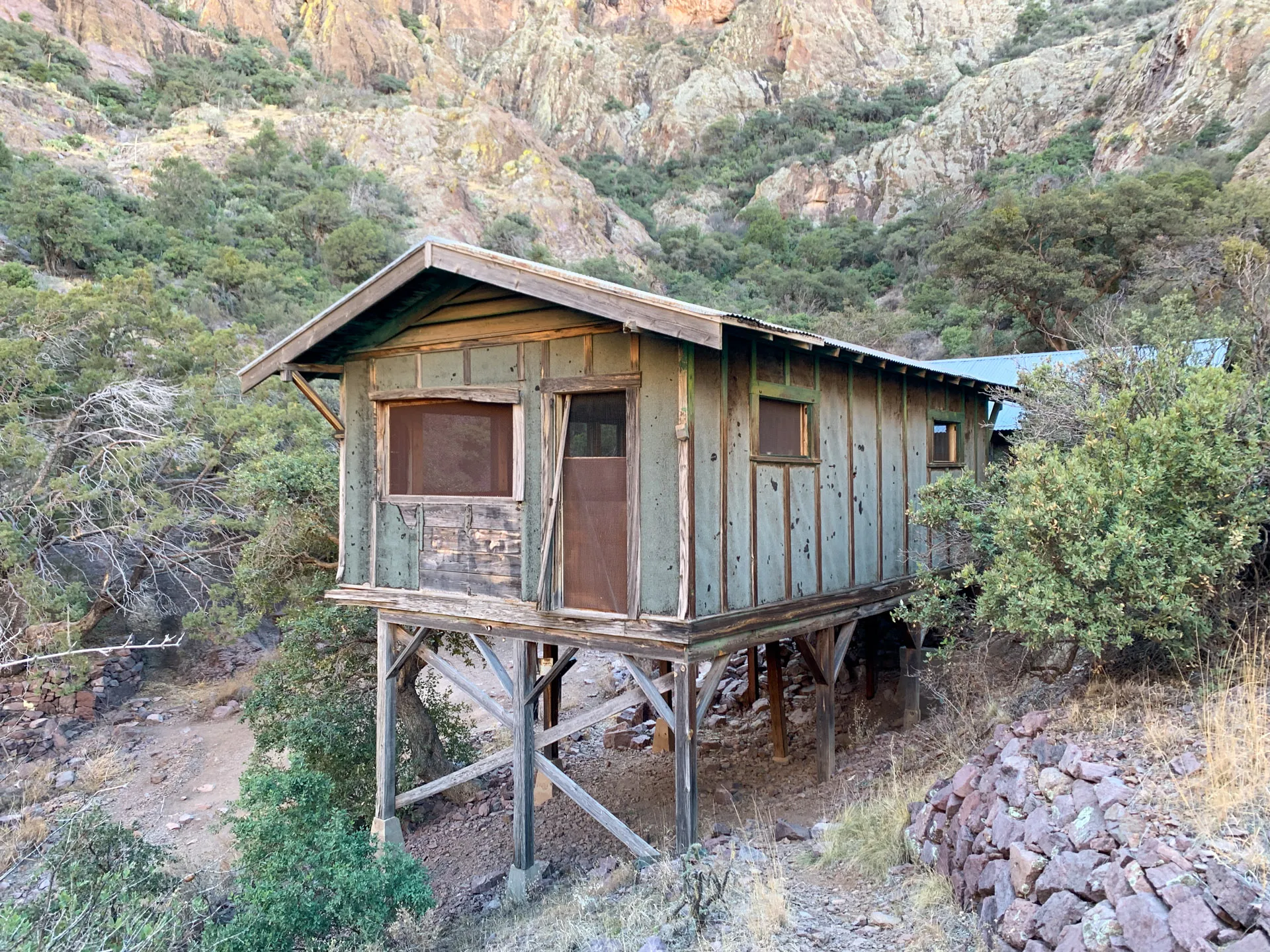 A weathered wooden cabin stands on stilts amidst rugged terrain. The structure has green and brown siding, with a few windows and a small porch. It's surrounded by dense greenery and set against a backdrop of steep, rocky cliffs under a clear sky.