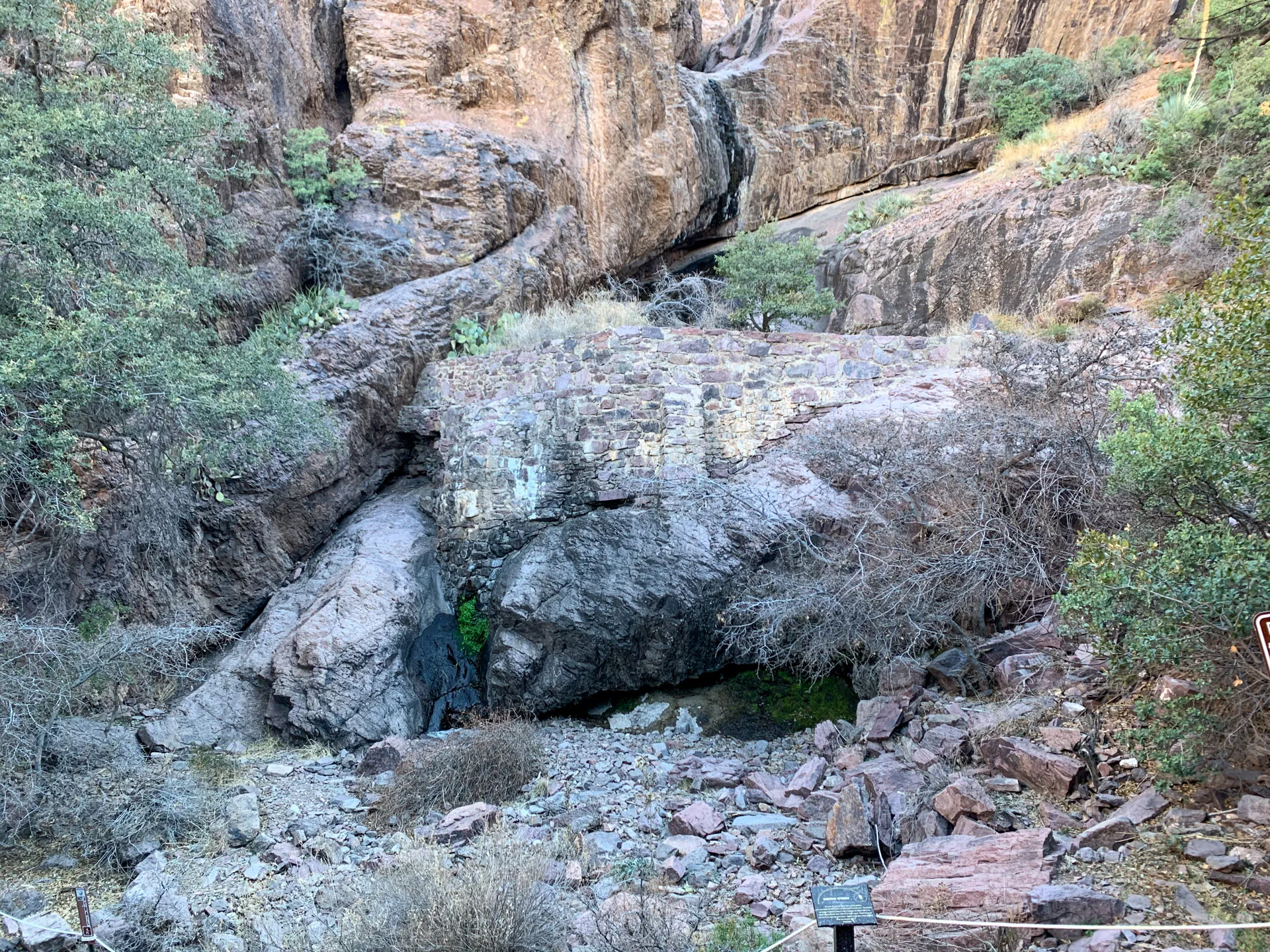 Rocky terrain with a small waterfall cascading down layered stone. Sparse green shrubs and trees dot the landscape amidst dry, rugged rocks. A wire fence and a small informational sign are visible in the foreground. The scene is set against a backdrop of tall rock formations.
