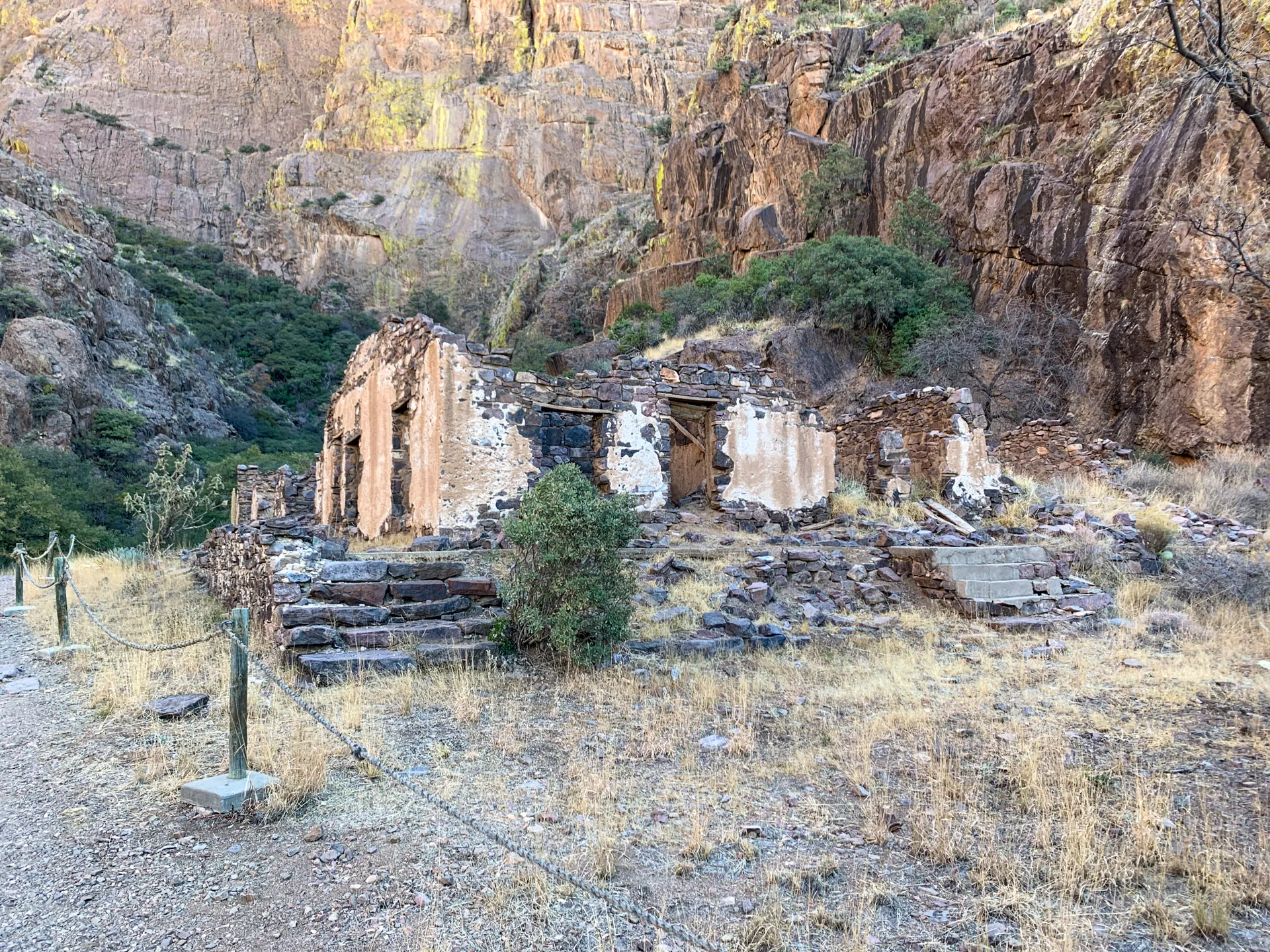 Ruins of a stone building with crumbling walls and no roof, surrounded by dry grass and rugged terrain. A rope fence leads to the structure. Rocky cliffs and sparse vegetation are in the background, under a clear blue sky. A few steps are visible in front of the ruins.