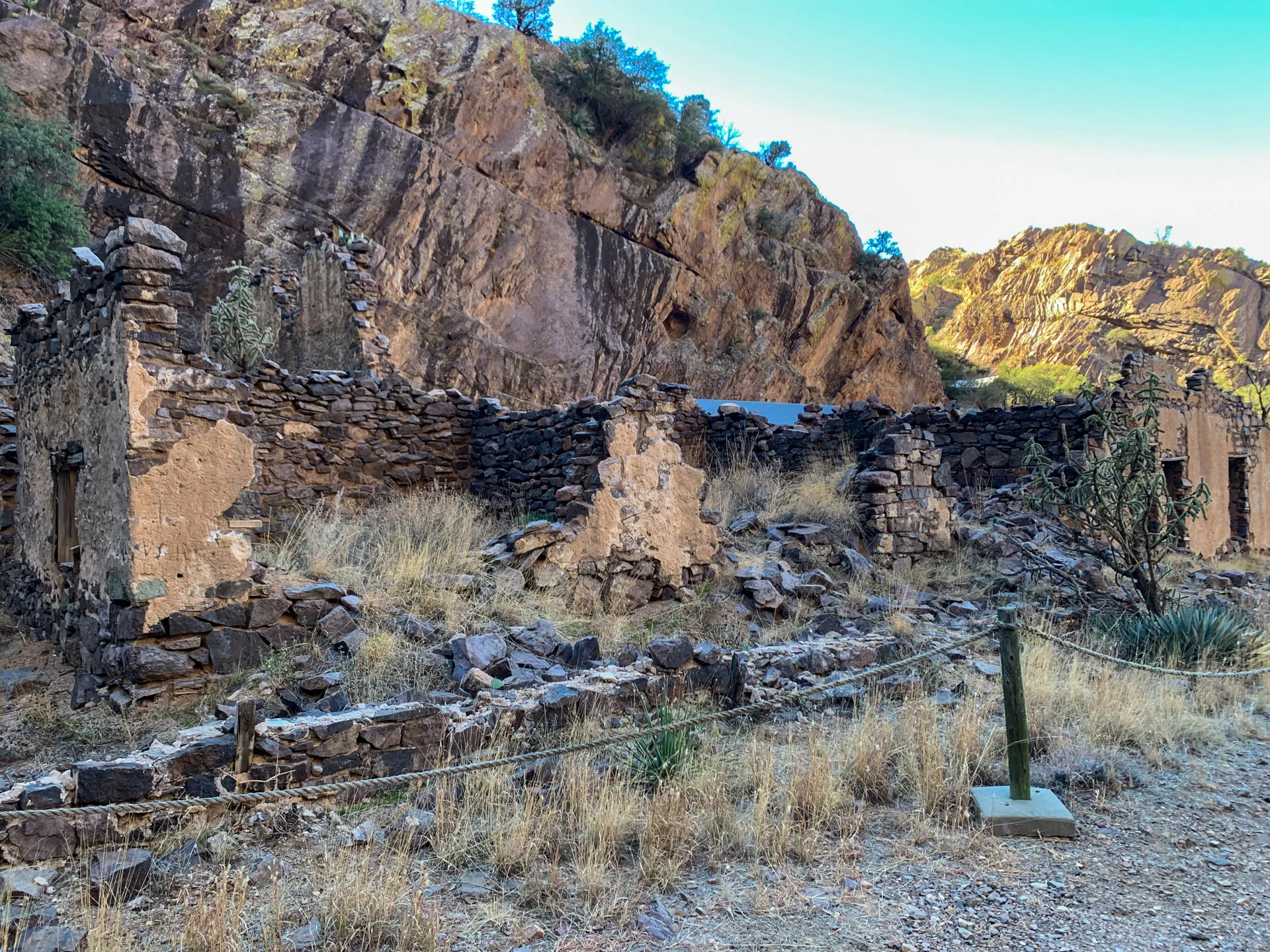 Ruins of a stone building with crumbling walls stand amid dry grass and rocky terrain. A rope barrier surrounds the site. In the background, steep, sunlit cliffs rise against a clear blue sky, while scattered vegetation grows along the rocky surfaces.