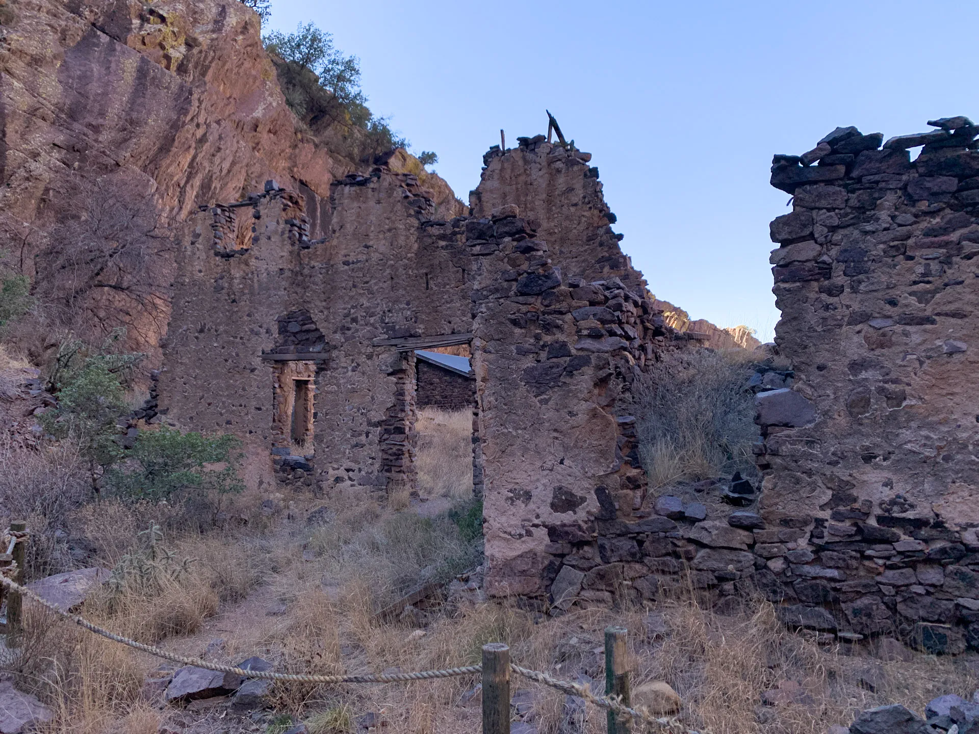 Ruins of a stone building against a rocky hillside. The structure's walls are partially standing, with visible door and window openings. Dry grass and a rope barrier are in the foreground, while a clear blue sky is above. Sparse greenery grows around the rocky landscape.