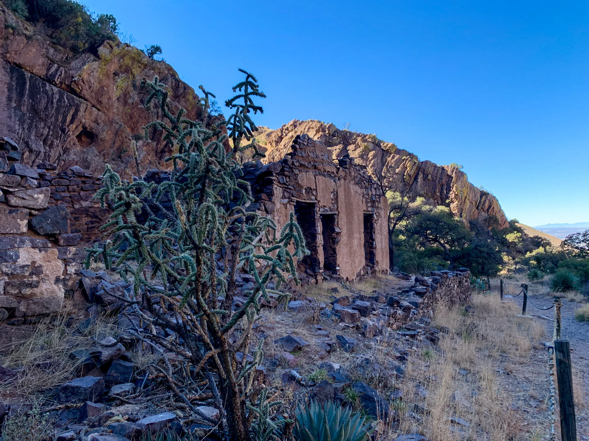 Desert landscape with an old, weathered stone building surrounded by rocky terrain. A cactus is in the foreground, and a rugged, sunlit mountain range is in the background. The sky is clear and blue, adding contrast to the earthy tones of the scene. Sparse vegetation dots the landscape.