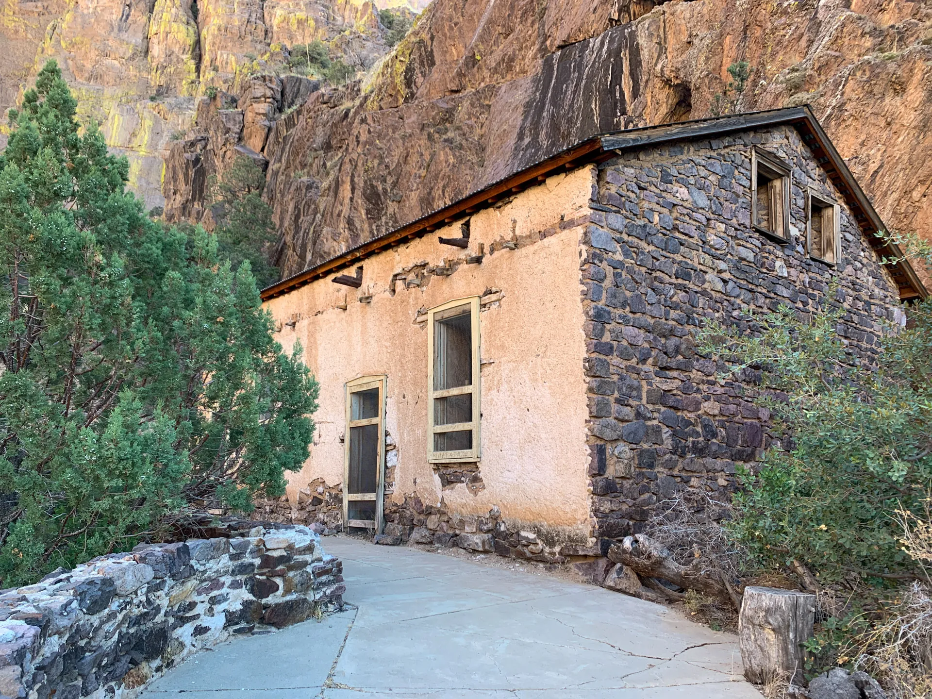 A rustic stone cabin with slightly weathered wooden features is set against a rugged cliff backdrop. The structure has two small windows near the roof and green shrubs in the foreground. Sunlight highlights the rocky landscape, creating a serene and historical ambiance.