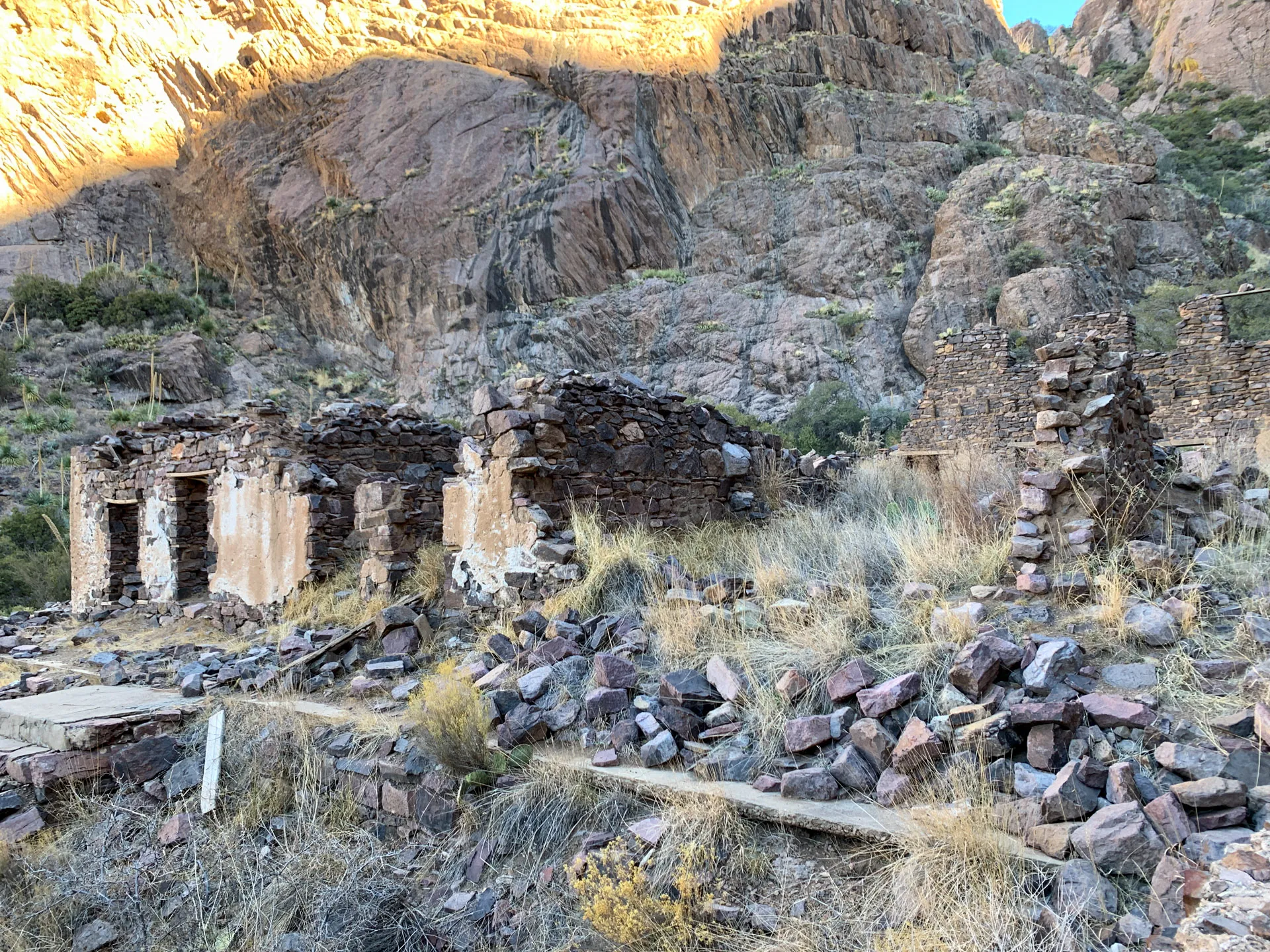 Ruins of an old stone building are set against a rocky mountainside. The building's walls are partially intact, with some sections collapsed. The surrounding area is scattered with rocks and dry grass, and the mountain is illuminated by sunlight, casting shadows on the terrain.