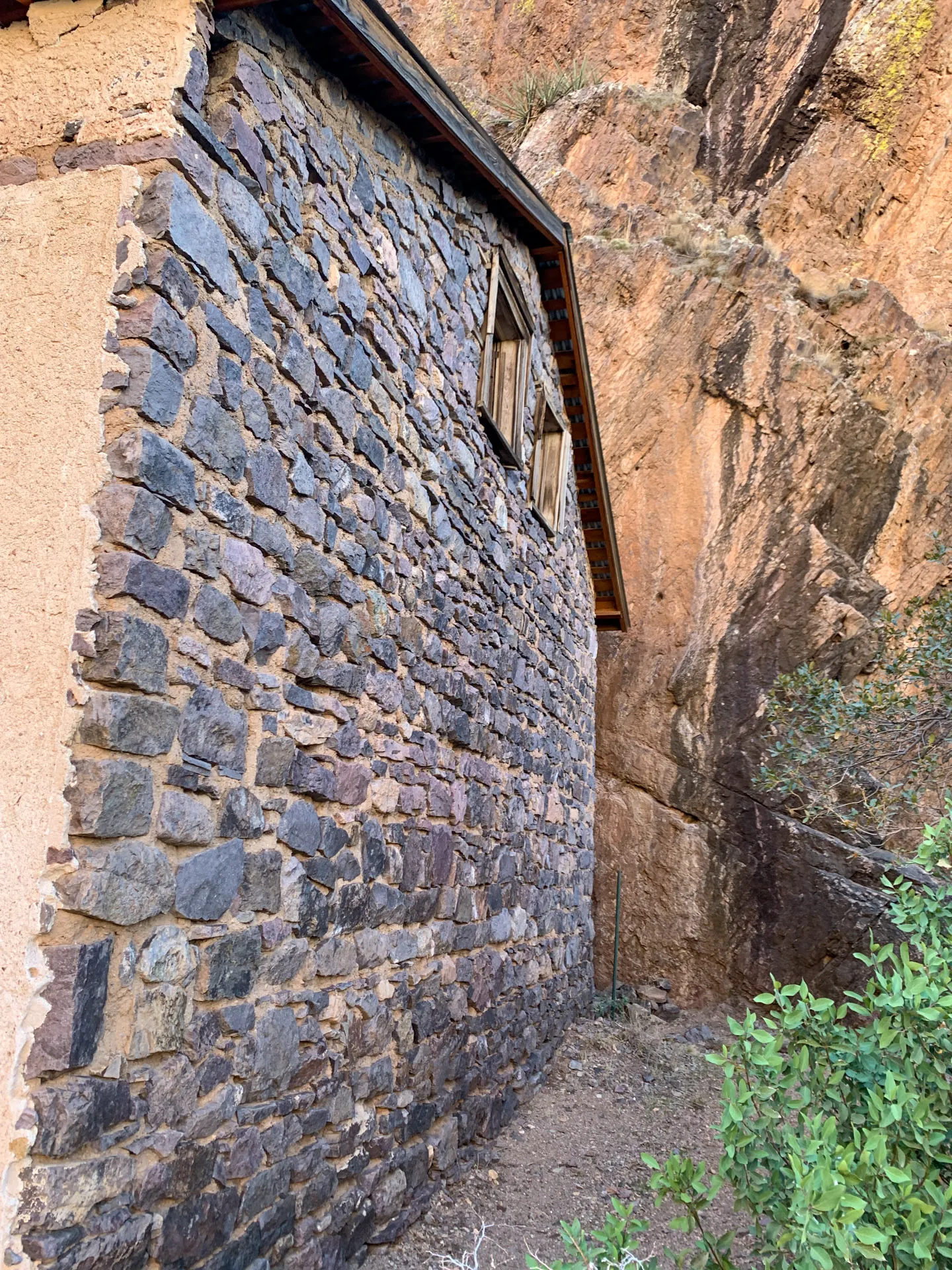 Stone building with two wooden windows set into a rocky cliff. The building's walls are made of irregular stones, and its roof is tiled. Vegetation grows at the base, and the backdrop features brown, rugged rock formations. The scene is outdoors on a sunny day.