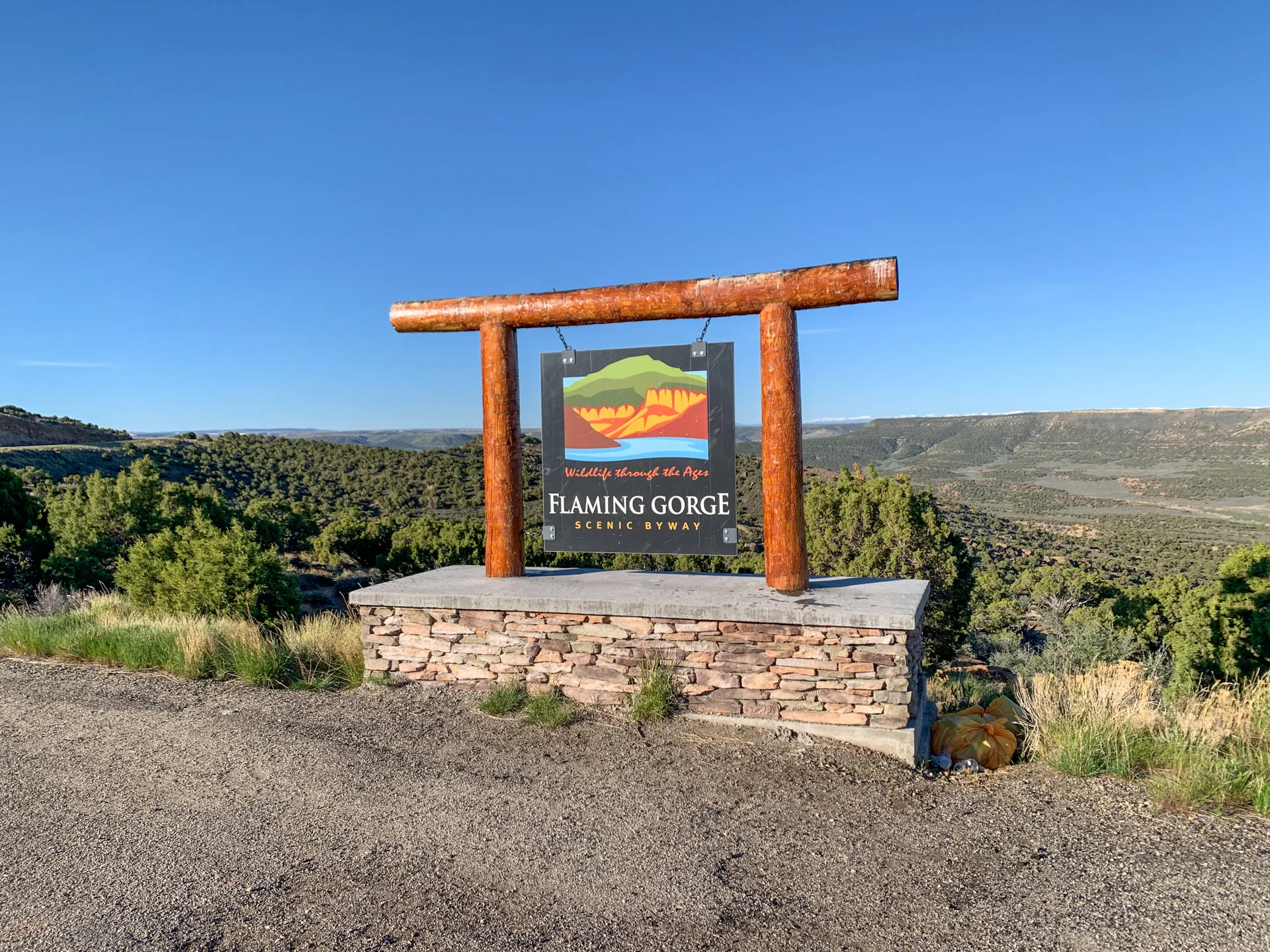 A wooden and stone sign reading "Flaming Gorge Scenic Byway" under a logo showing a stylized landscape is set beside a road. Behind it, a vast view of the Flaming Gorge area with lush greenery and hills under a clear blue sky is visible.