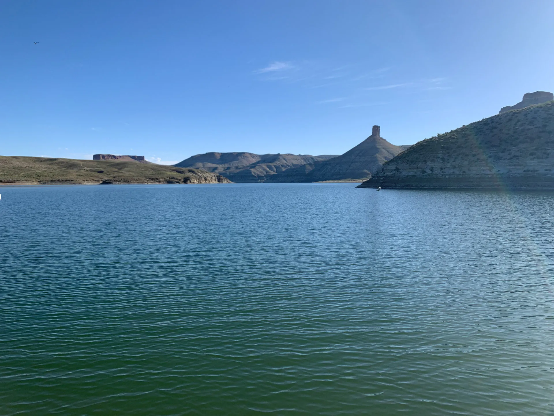 A serene lake with calm blue water and distant hills under a clear blue sky. The landscape features a prominent hill with a distinctive peak on the right. Sparse vegetation covers the hills, and the sun illuminates the scene, creating a peaceful, natural setting.