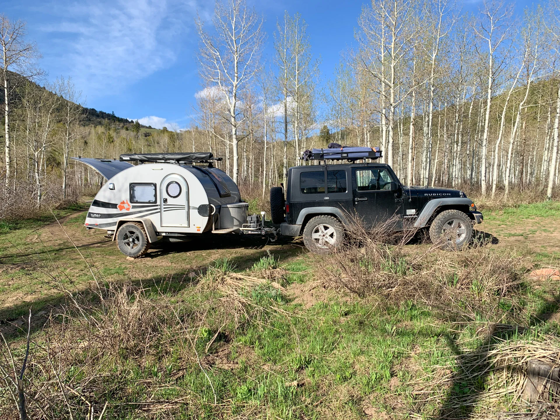 A black off-road vehicle is parked on a grassy field, towing a small teardrop camper. The scene is set in a natural environment with sparse trees and hills in the background under a clear blue sky. The camper has a rounded shape and features a logo on its side.