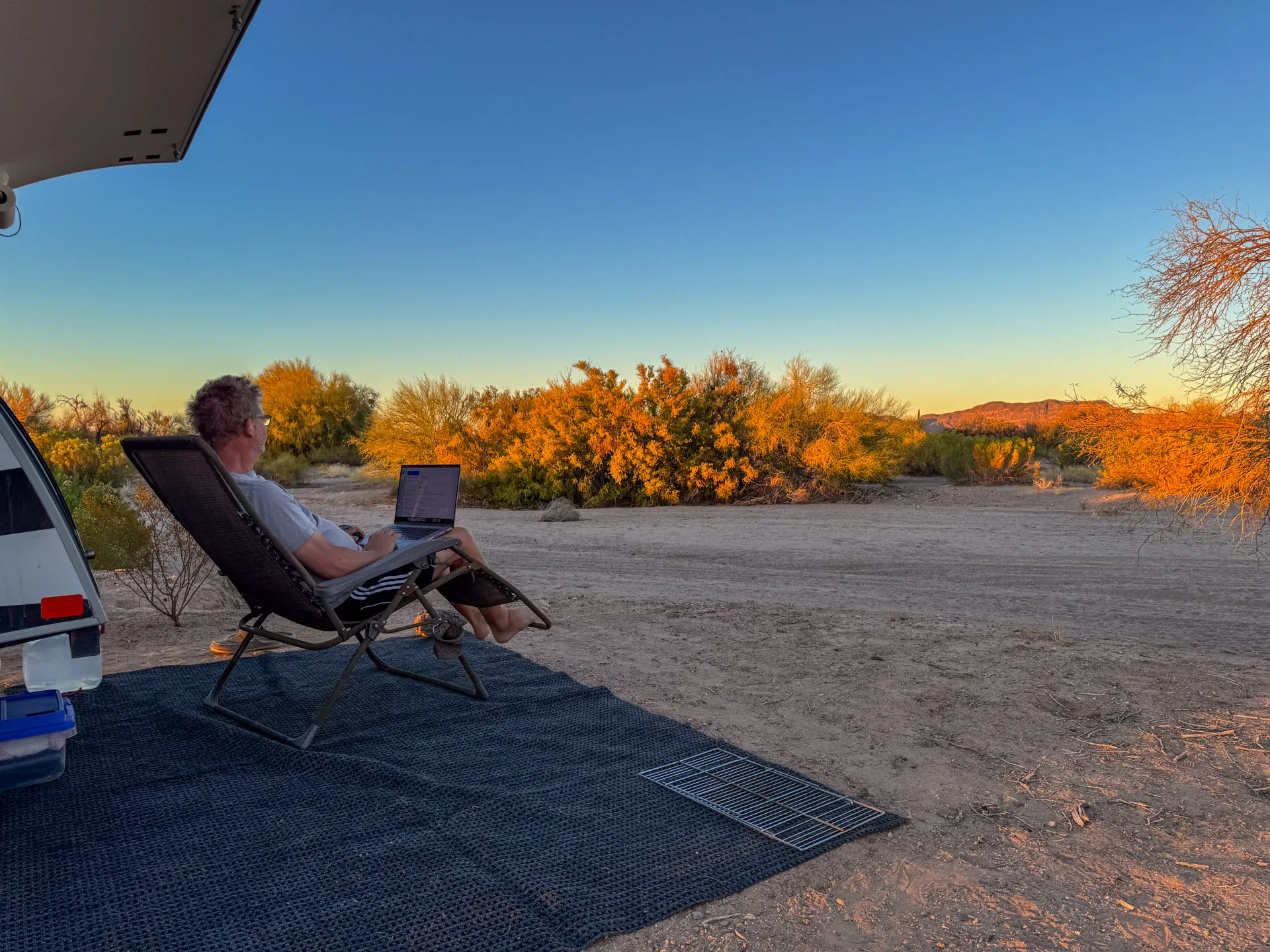 A person is sitting on a reclining chair under an RV awning in a desert landscape. They are using a laptop on their lap, facing towards the setting sun. The surroundings include sparse shrubs and distant low hills, all bathed in warm, golden light. The ground is sandy, covered with a mat.
