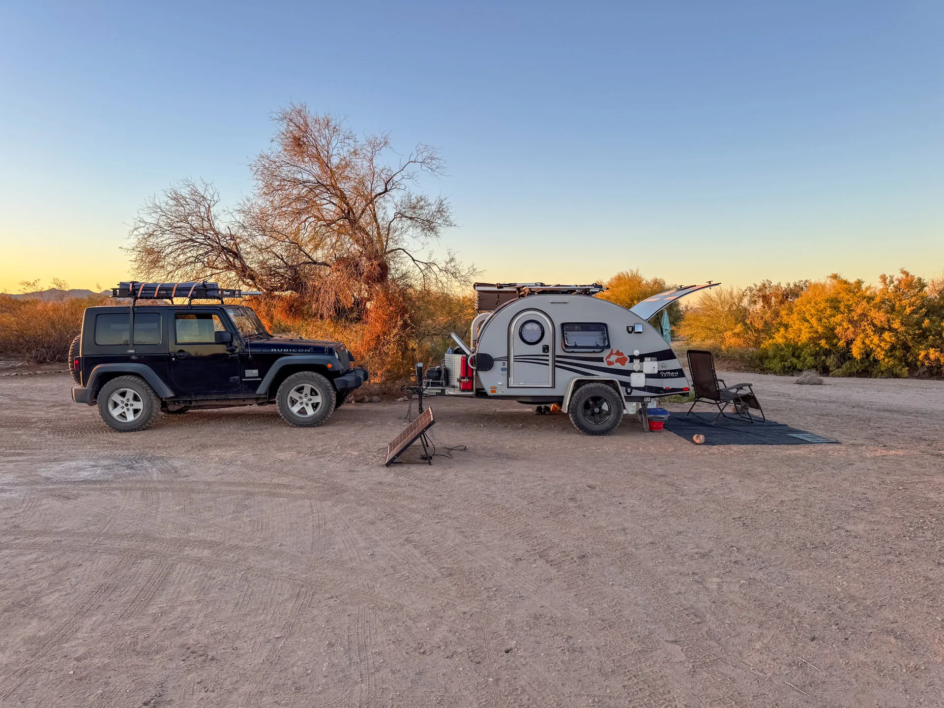 A compact camping scene on a clear day with a blue sky. A dark blue Jeep is parked next to a small white teardrop trailer. The trailer's door and storage compartments are open. Sparse desert vegetation and a leafless tree are in the background, while camping equipment is set up outside.