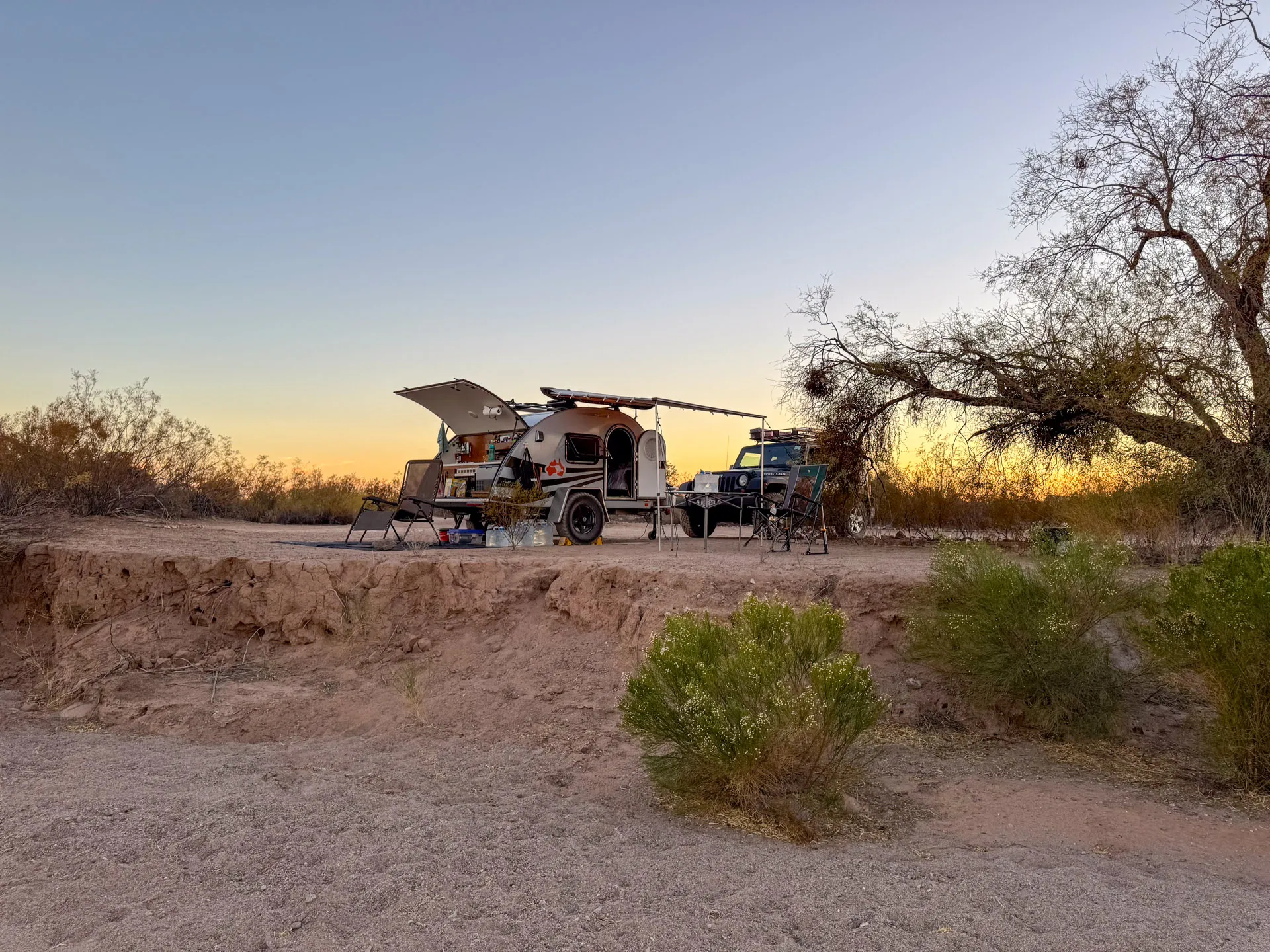 A teardrop camper and pickup truck parked at a sandy, elevated campsite during sunset. The camper's hatch is open, revealing camping supplies. Sparse desert plants and shrubs are in the foreground, with bare trees and a clear sky in the background.