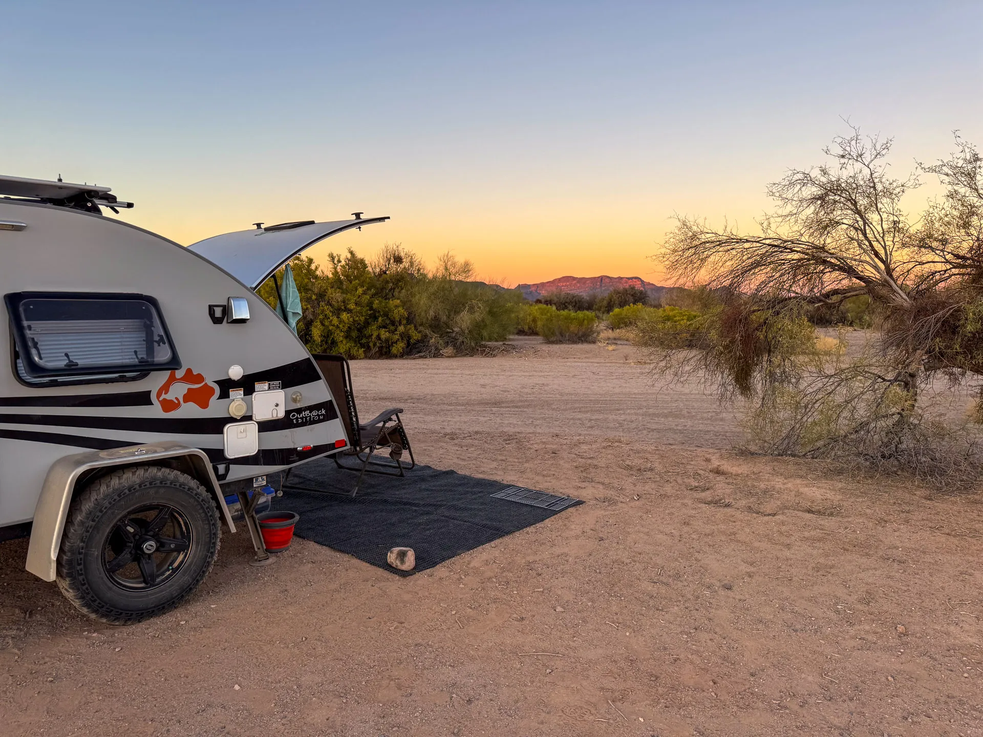 A small teardrop camper is parked on a desert landscape at sunset. The camper has its hatch open, revealing a cozy interior. A black mat, a water bowl, and a red bucket sit on the ground. Sparse desert vegetation surrounds the area, with hills visible in the distance under a colorful sky.