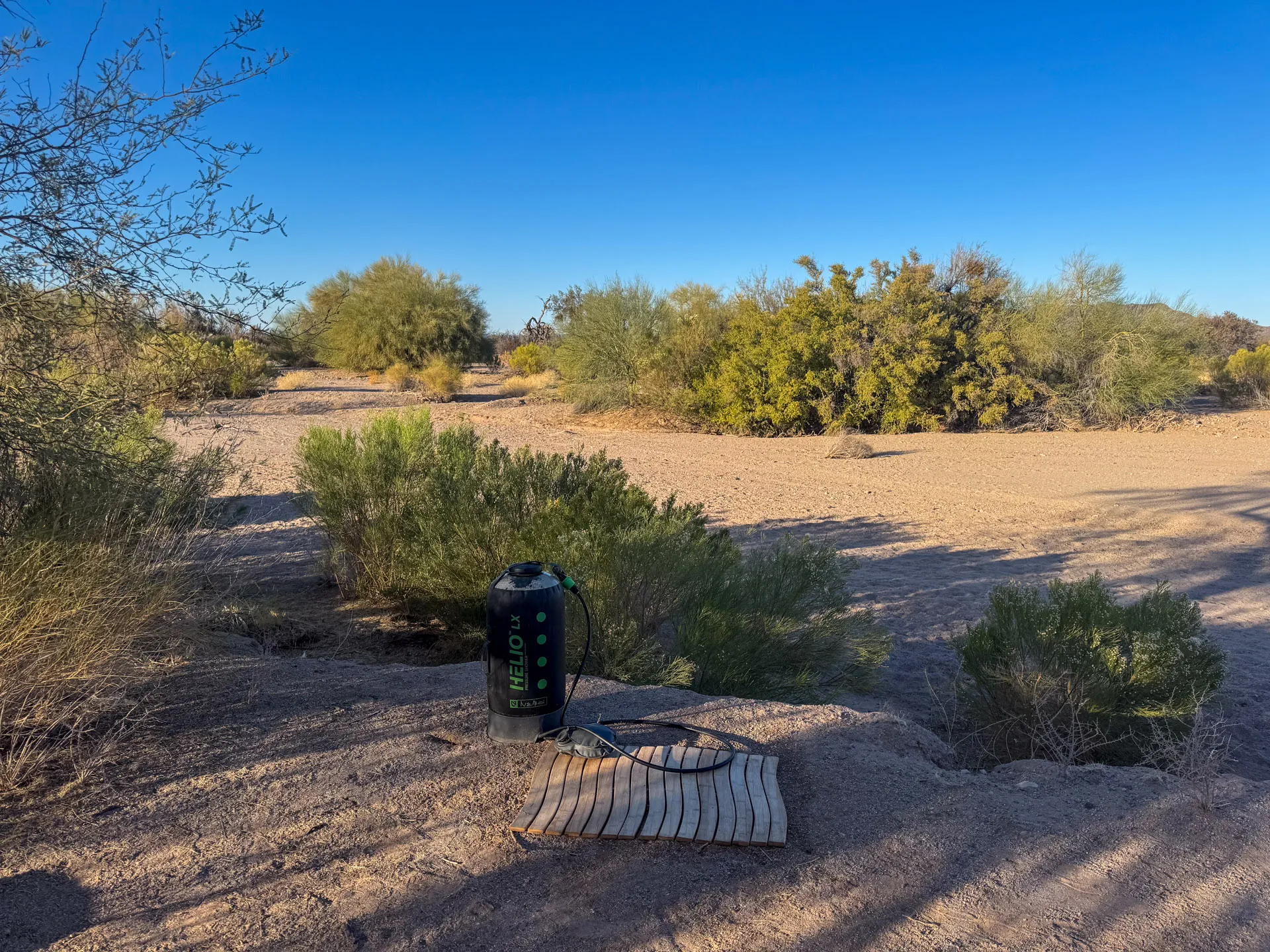 A desert landscape under a clear blue sky features scattered bushes and small trees. In the foreground, a solar shower sits on a patch of sandy soil next to a folded wooden mat. Shadows of nearby plants stretch across the ground, indicating early or late in the day.
