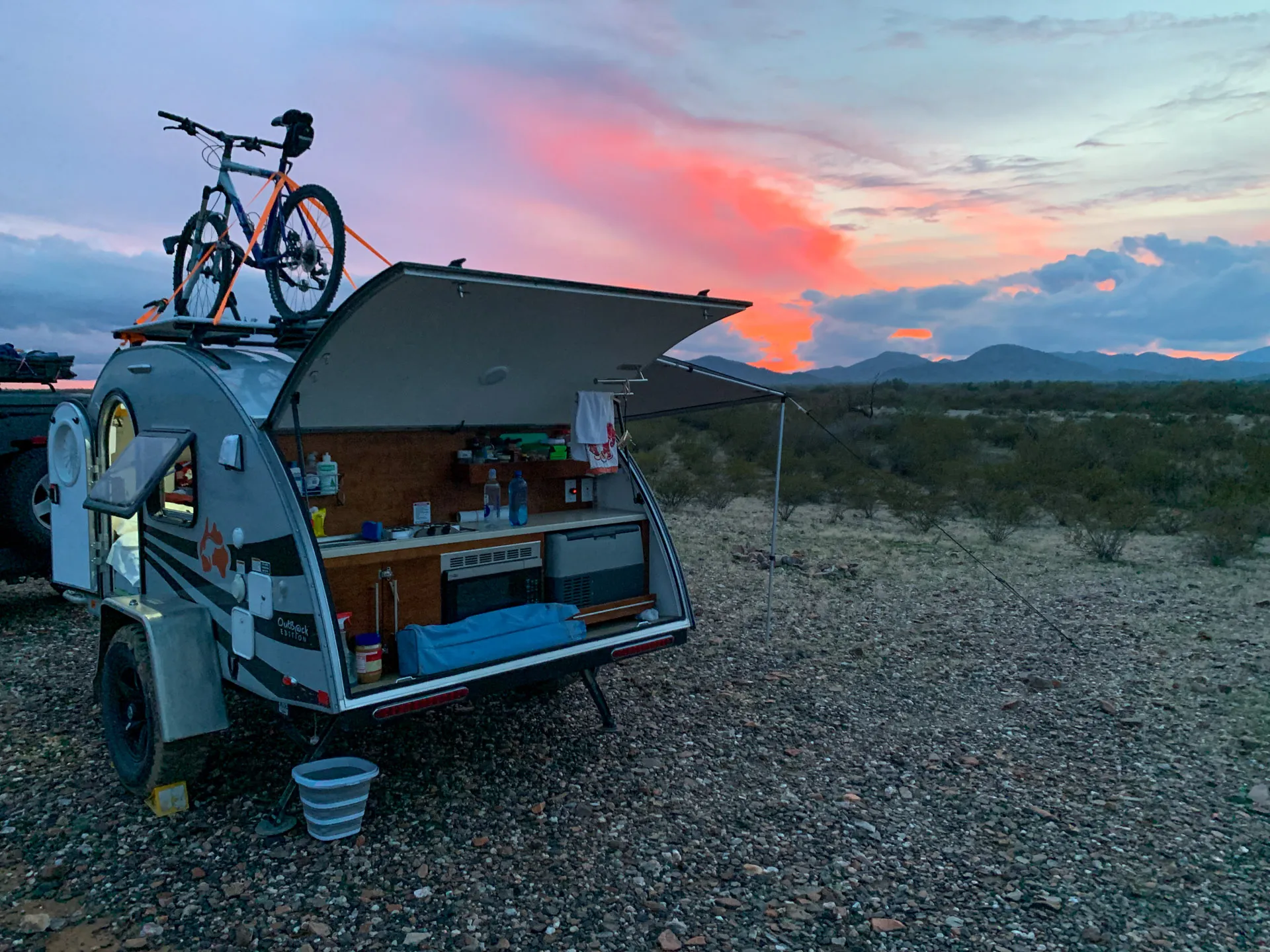 Small teardrop camper with outdoor kitchenette open, displaying storage and cooler. A bicycle is mounted on the roof. The camper is parked on a rocky terrain with mountains in the distance. The sky features a colorful sunset with pink, orange, and blue hues.
