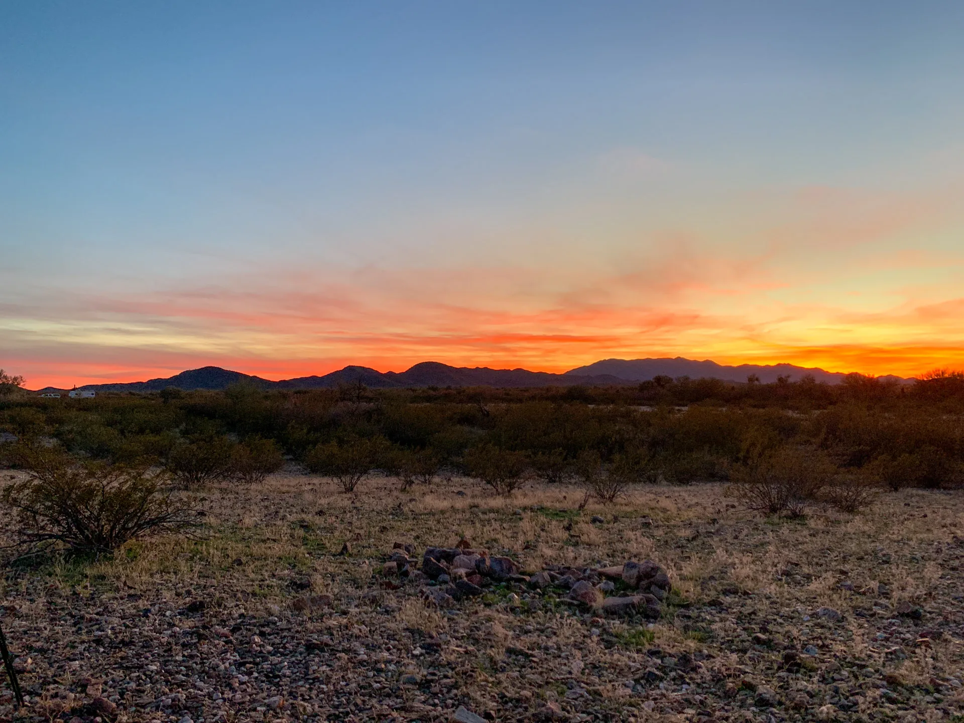 Sunset over a desert landscape with rocky foreground and sparse vegetation. The sky is a gradient of blue to orange, with the sun setting behind distant dark mountain silhouettes. A small pile of rocks is in the center of the image, adding texture to the arid scene.