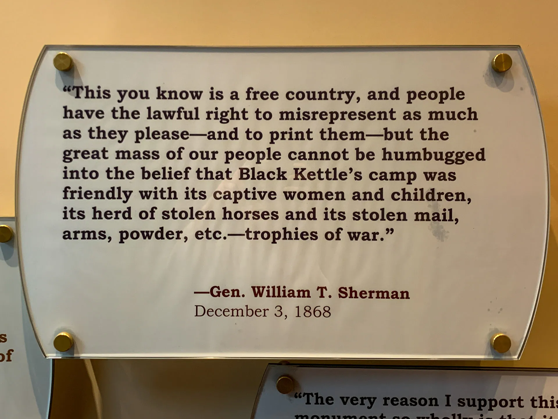 A plaque displays a quote by Gen. William T. Sherman dated December 3, 1868. It discusses freedom in the U.S. and refutes claims of kindness toward Black Kettle’s camp, mentioning captives and stolen items described as “trophies of war.” The plaque is mounted on a wall with gold screws.