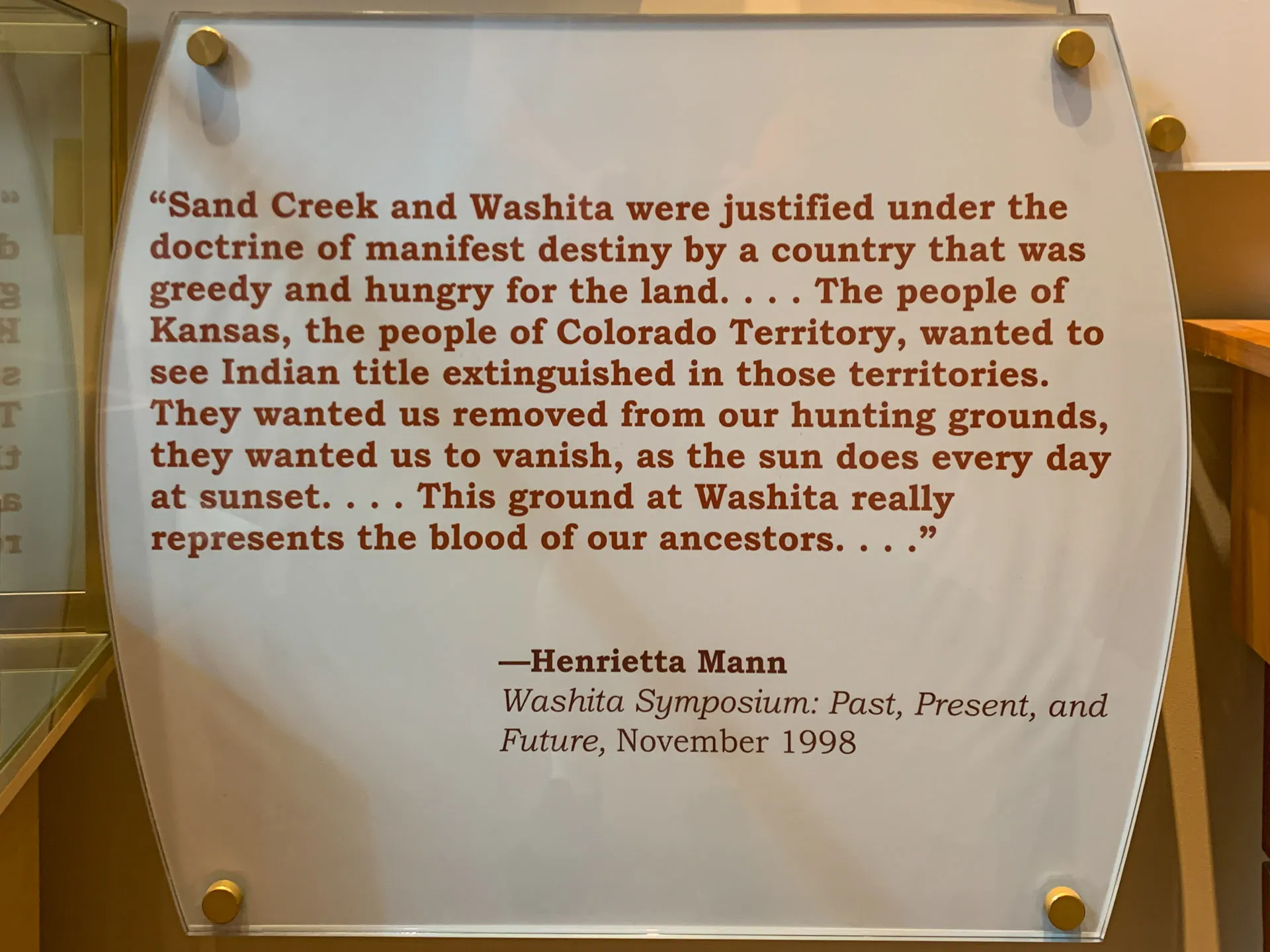 A display case with a quote by Henrietta Mann from the "Washita Symposium: Past, Present, and Future," November 1998. The quote discusses the historical impact of the doctrine of manifest destiny and its effects on indigenous lands and cultures, highlighting the loss faced by Native Americans.