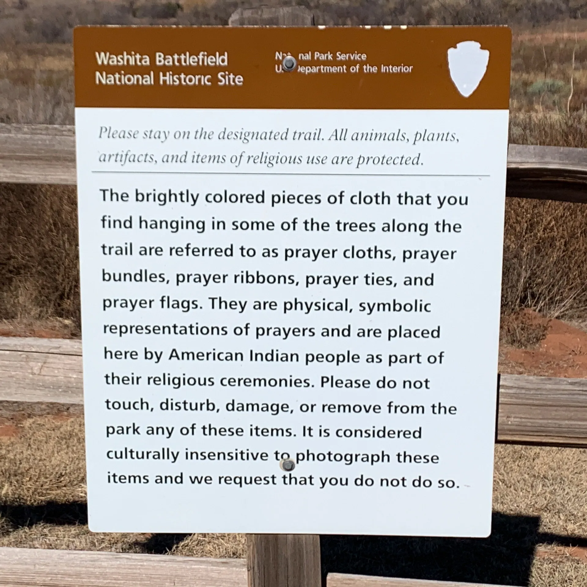 Sign at Washita Battlefield National Historic Site explaining that brightly colored cloth pieces on trees are prayer items used by American Indian people. They symbolize prayers and are protected. The sign advises against touching, disturbing, or photographing them.