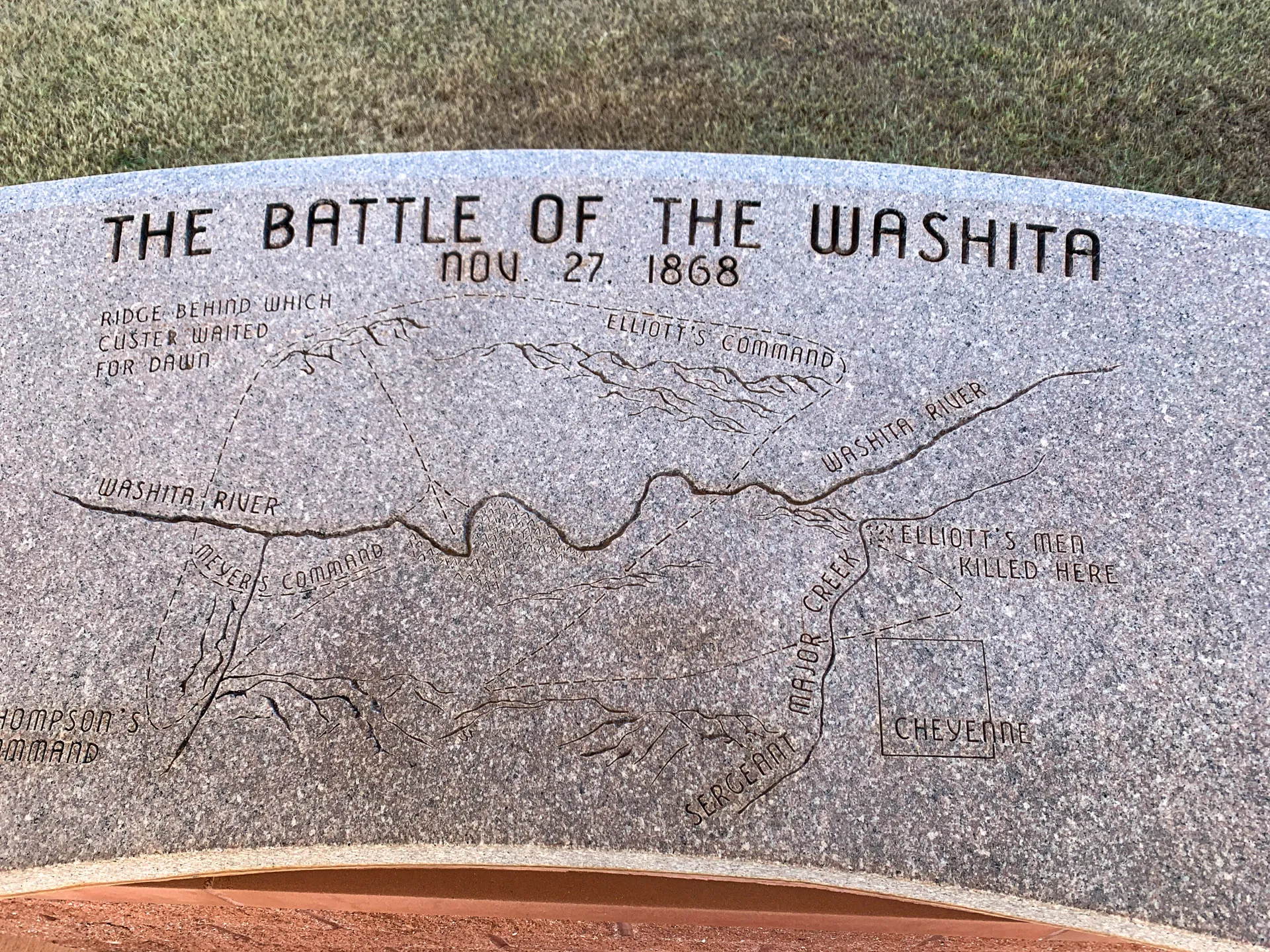 Stone monument depicting a map with "The Battle of the Washita, Nov 27, 1868" engraved. It shows key locations like the Washita River, Elliott's Command, Major Elliot's group, and Cheyenne. The map includes topographical features and notes on troop positions, with grass in the background.