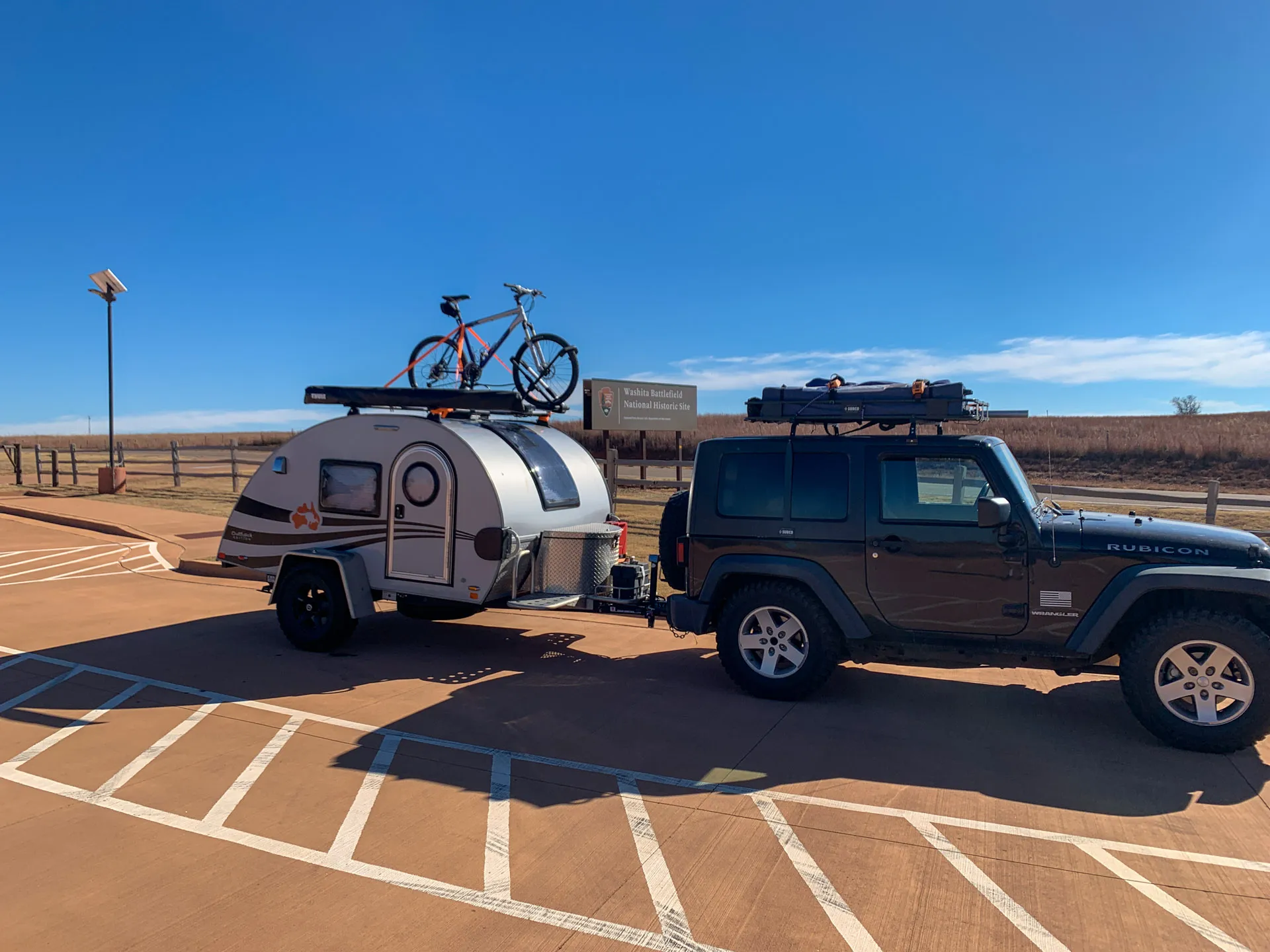 A black Jeep with a roof rack is towing a small teardrop trailer in a parking lot. The trailer has a bicycle mounted on top. The scene is under a clear blue sky, with open fields and a sign in the background indicating the Washita Battlefield National Historic Site.