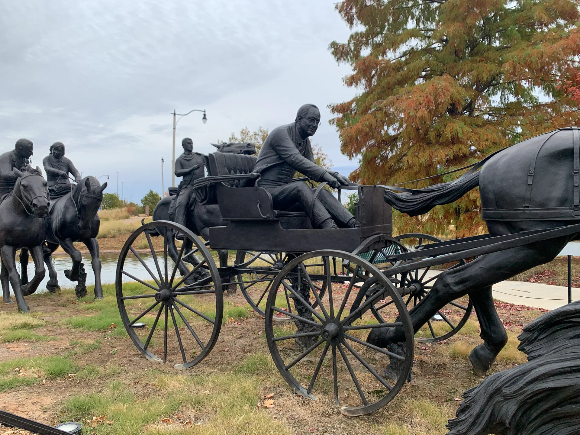 Bronze sculpture of a horse-drawn carriage with a driver holding reins, surrounded by other figures on horseback. The scene is set outdoors with a backdrop of trees and a cloudy sky. The artwork captures a historical moment in a realistic style.