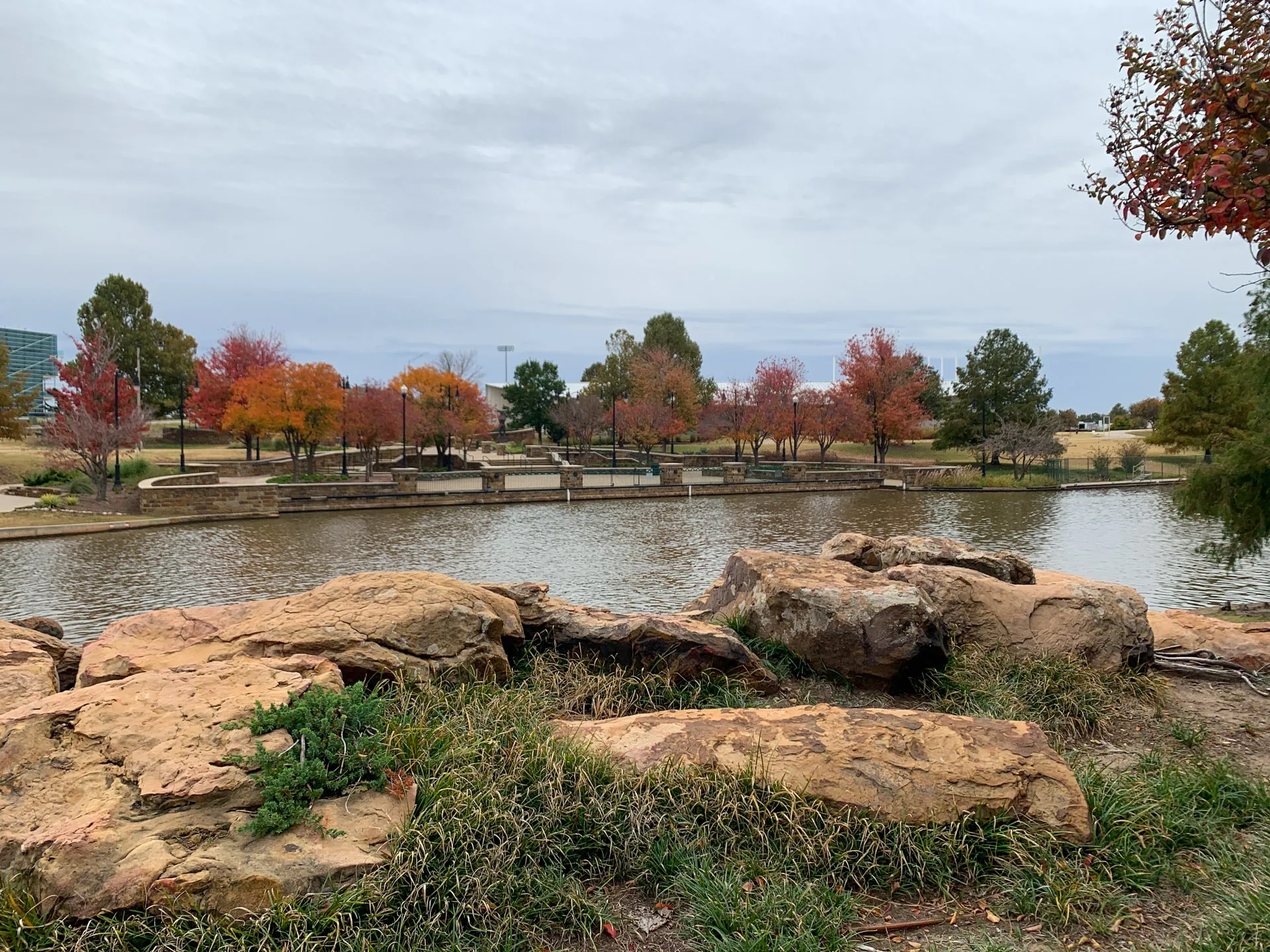 A tranquil park scene features a pond surrounded by large rocks and grassy areas in the foreground. Trees with autumn foliage in shades of red, orange, and green line the path across the pond. A cloudy sky adds a soft, muted backdrop to the landscape.