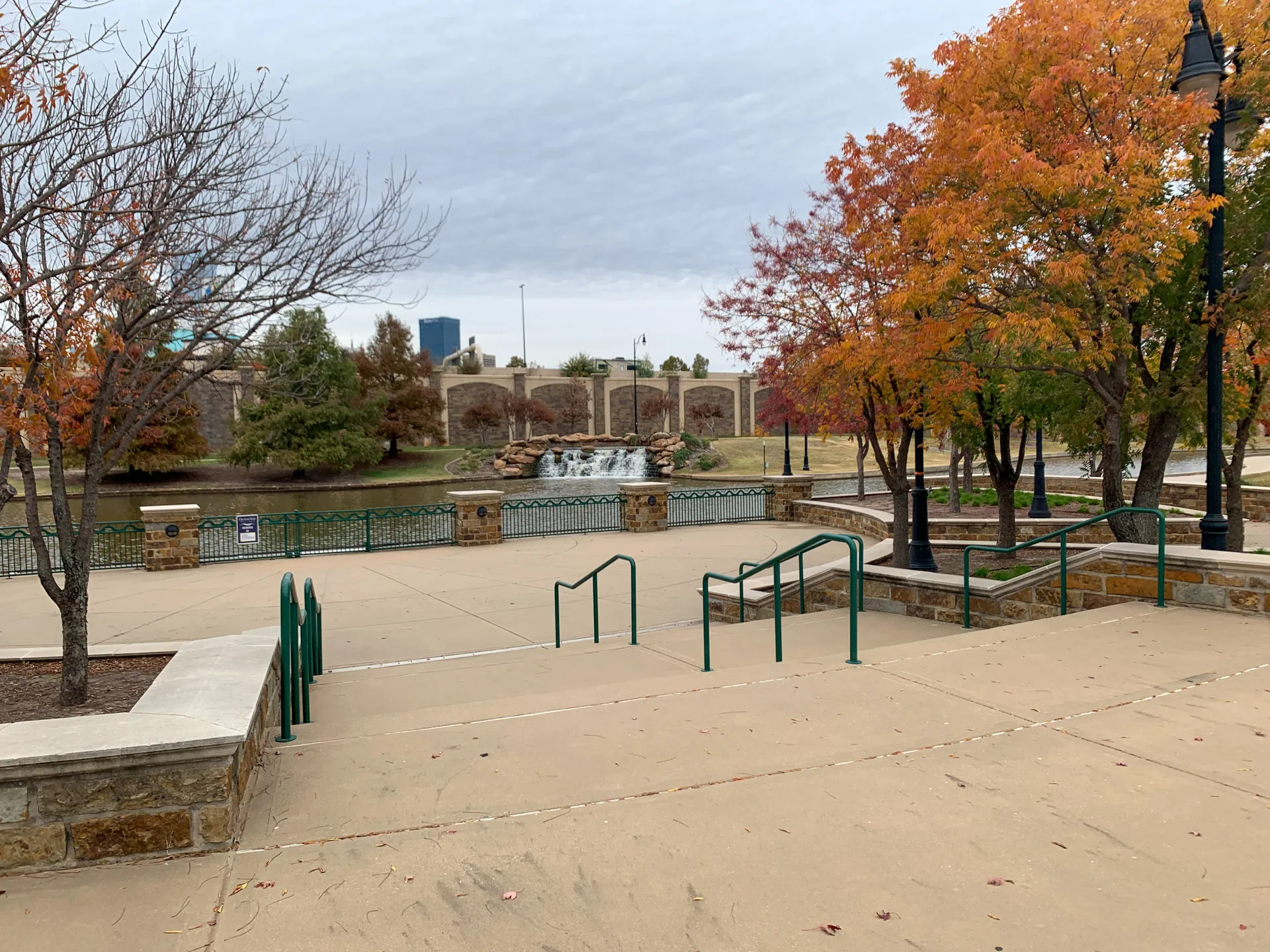 Park scene with paved walkway and green railings. A water feature with small waterfalls is in the background, surrounded by grassy hills and a stone wall. Trees with autumn foliage line the pathway. A cloudy sky is overhead, and city buildings can be seen in the distance.