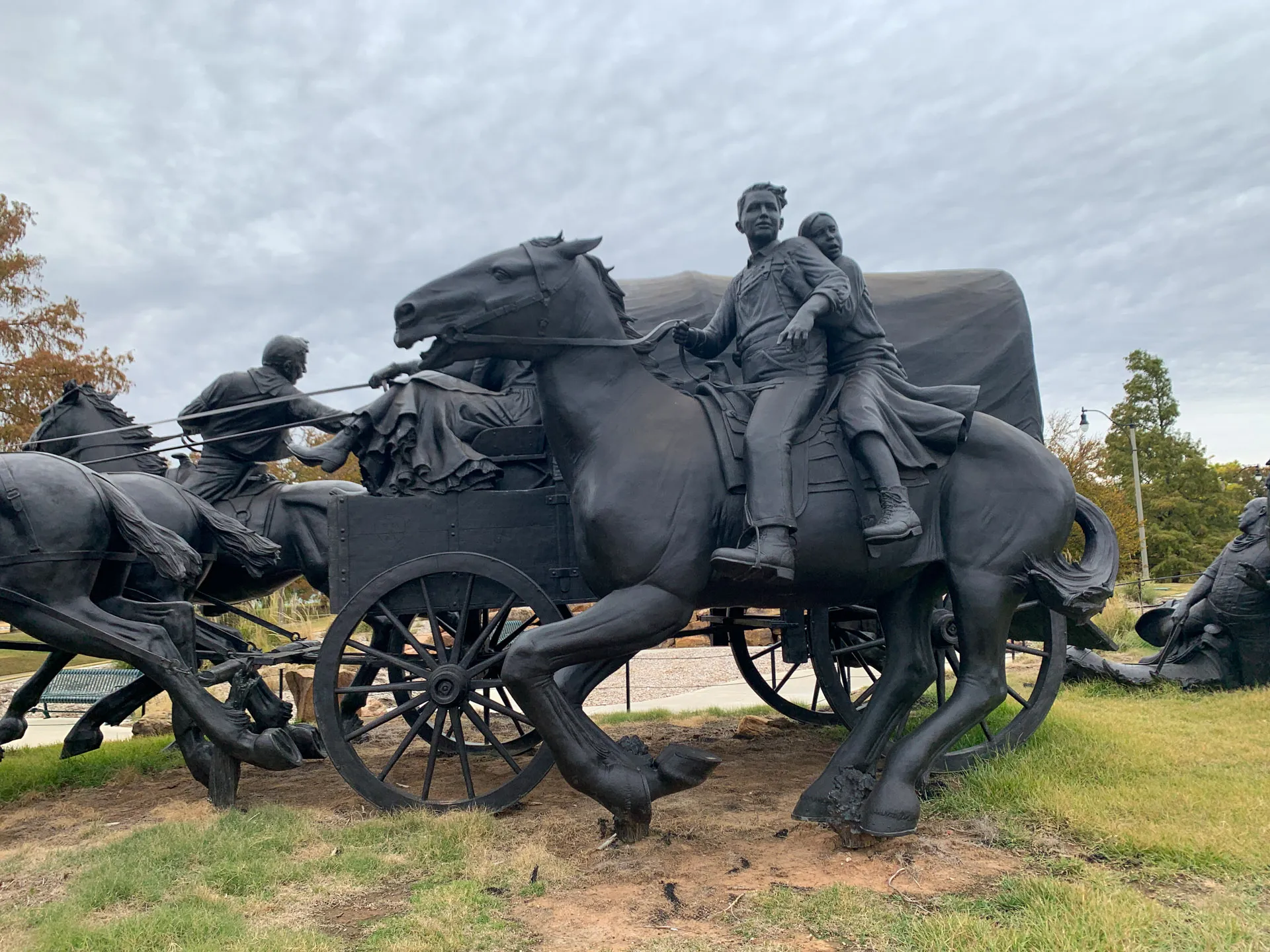 Bronze sculpture depicting pioneers on horseback and in a covered wagon. A man and woman ride a galloping horse in the foreground. Another rider and wagon are visible behind. The sky is overcast, and grass surrounds the sculpture. Trees are in the background.