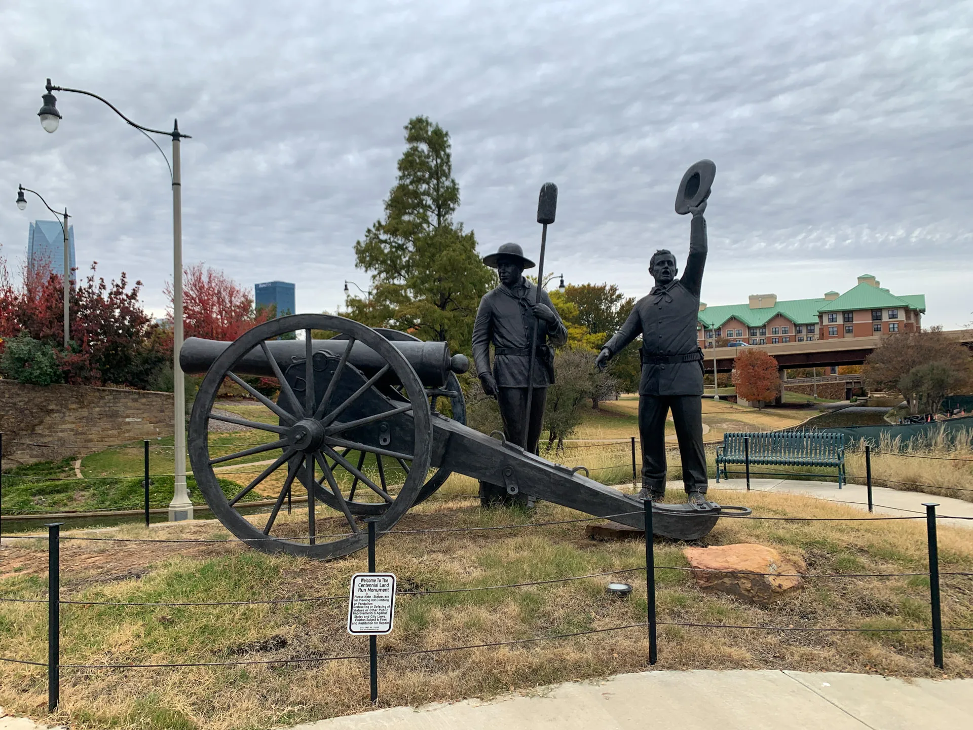 A bronze statue of two men stands next to a historic cannon. One man holds a hat aloft. The scene is in a grassy, fenced area with a cloudy sky above. Trees and a modern city skyline are visible in the background. A sign is placed near the statue on the grass.