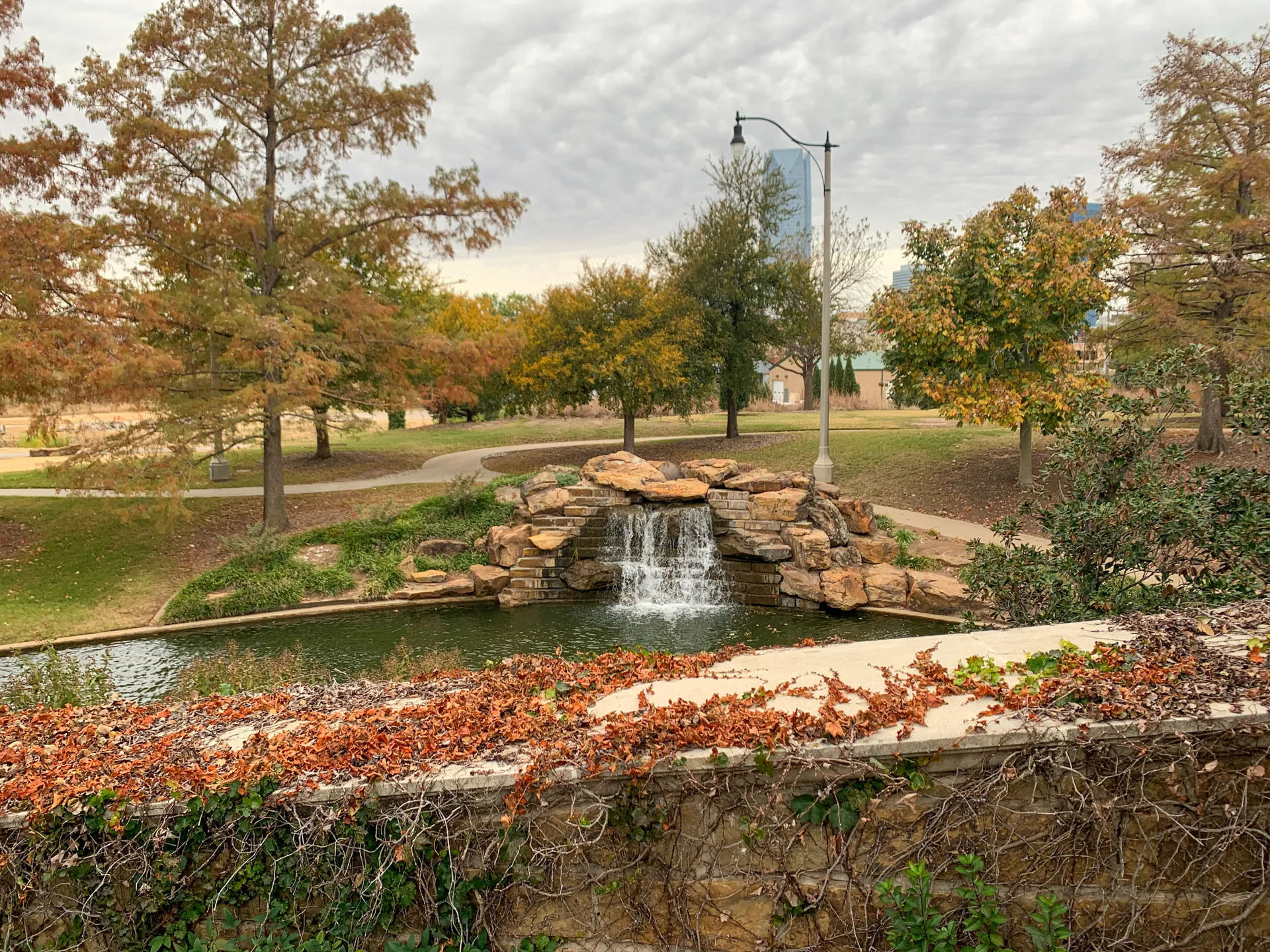 A serene park scene features a small waterfall cascading over a rock formation into a pond. Autumn trees with orange and yellow leaves line the background. A stone wall with ivy and red vines is in the foreground. A winding path and a tall lamppost are visible under a cloudy sky.