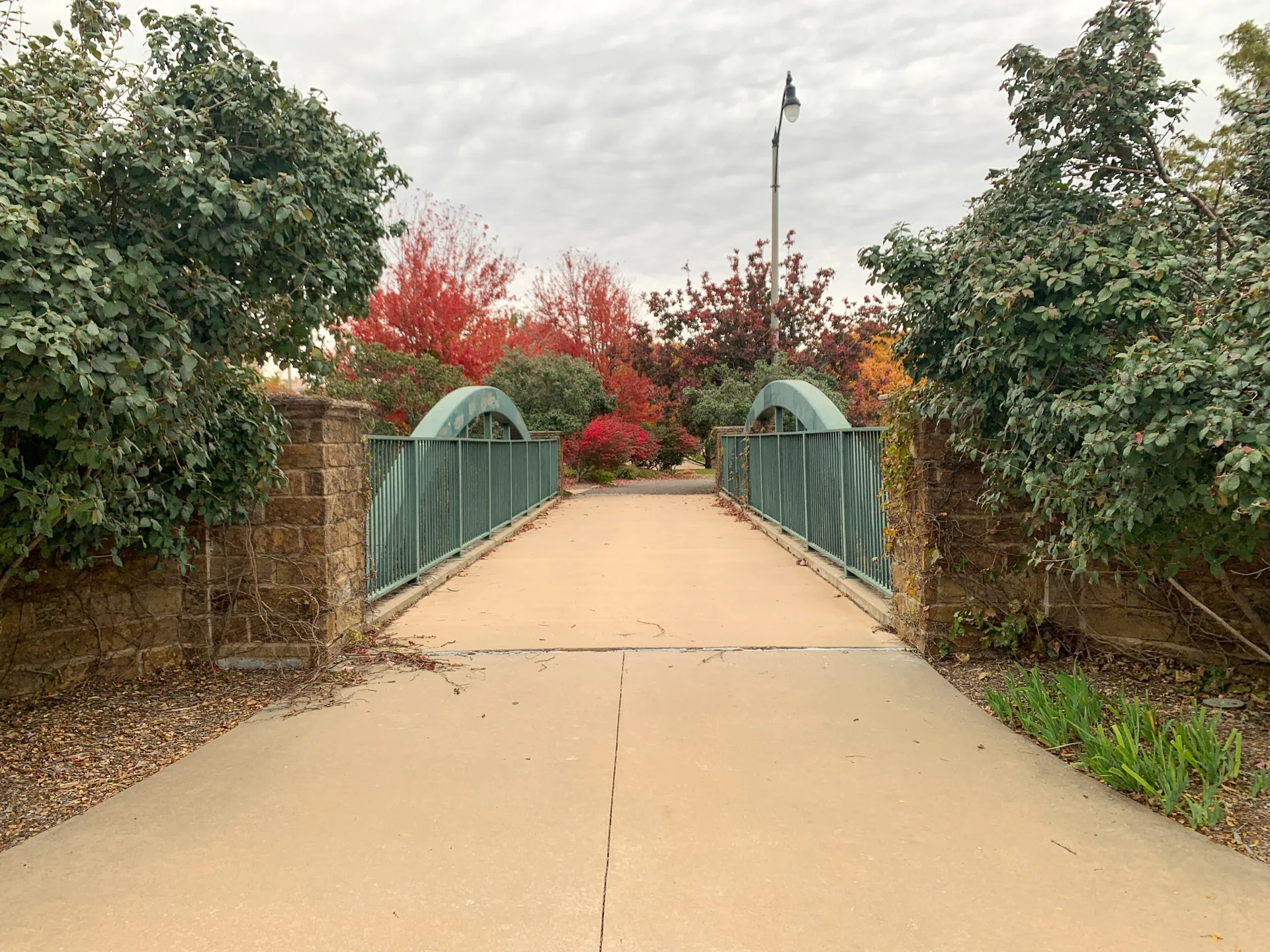 Pathway leading to a small green bridge flanked by stone pillars and metal railings. Surrounding trees display autumn foliage in shades of red and orange. Overcast sky and a streetlamp above the bridge enhance the scene's tranquil, seasonal atmosphere.